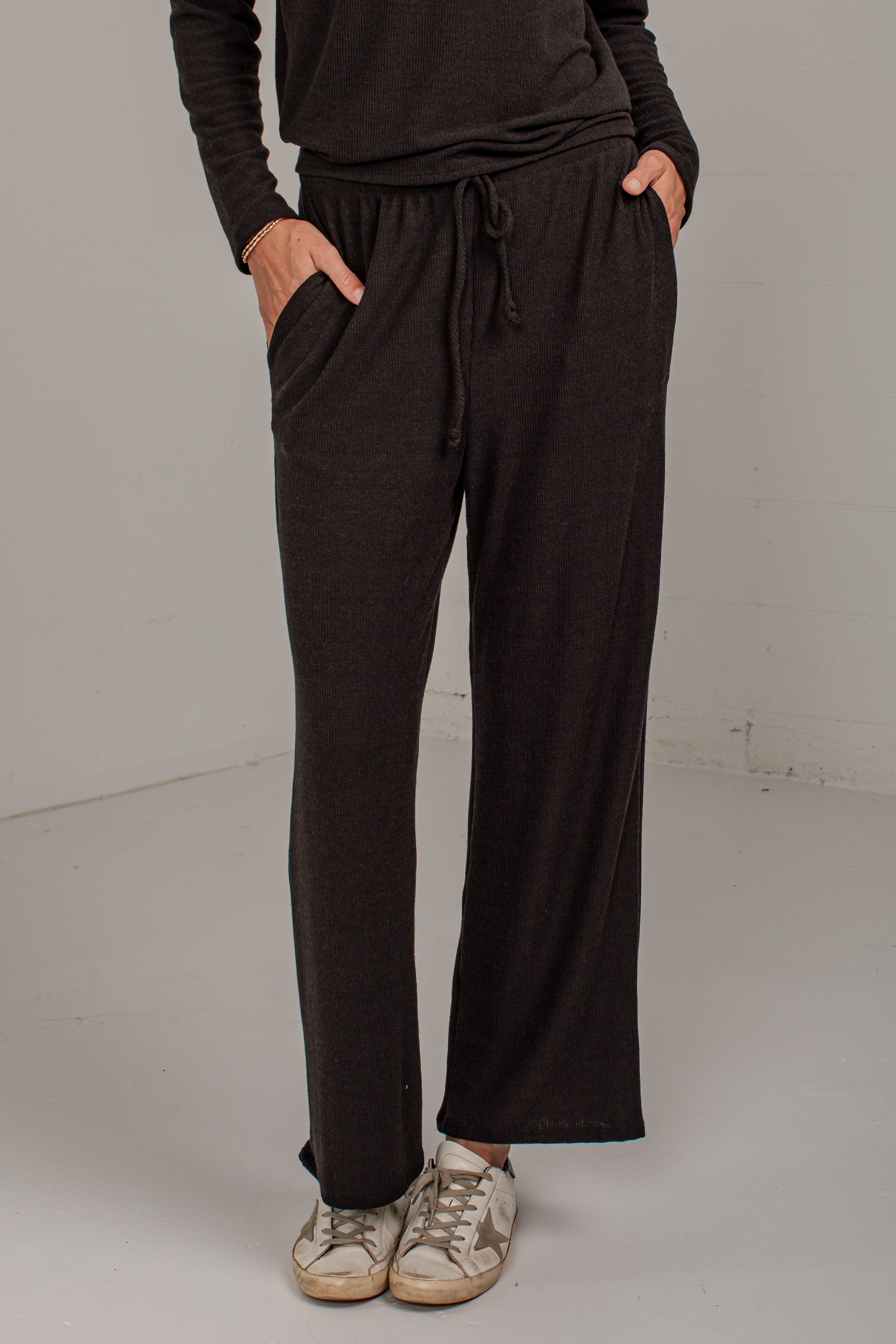 A person wearing dark brown wide-leg drawstring sweatpants with side pockets, paired with a matching dark top and beige sneakers, standing against a plain light gray studio background.