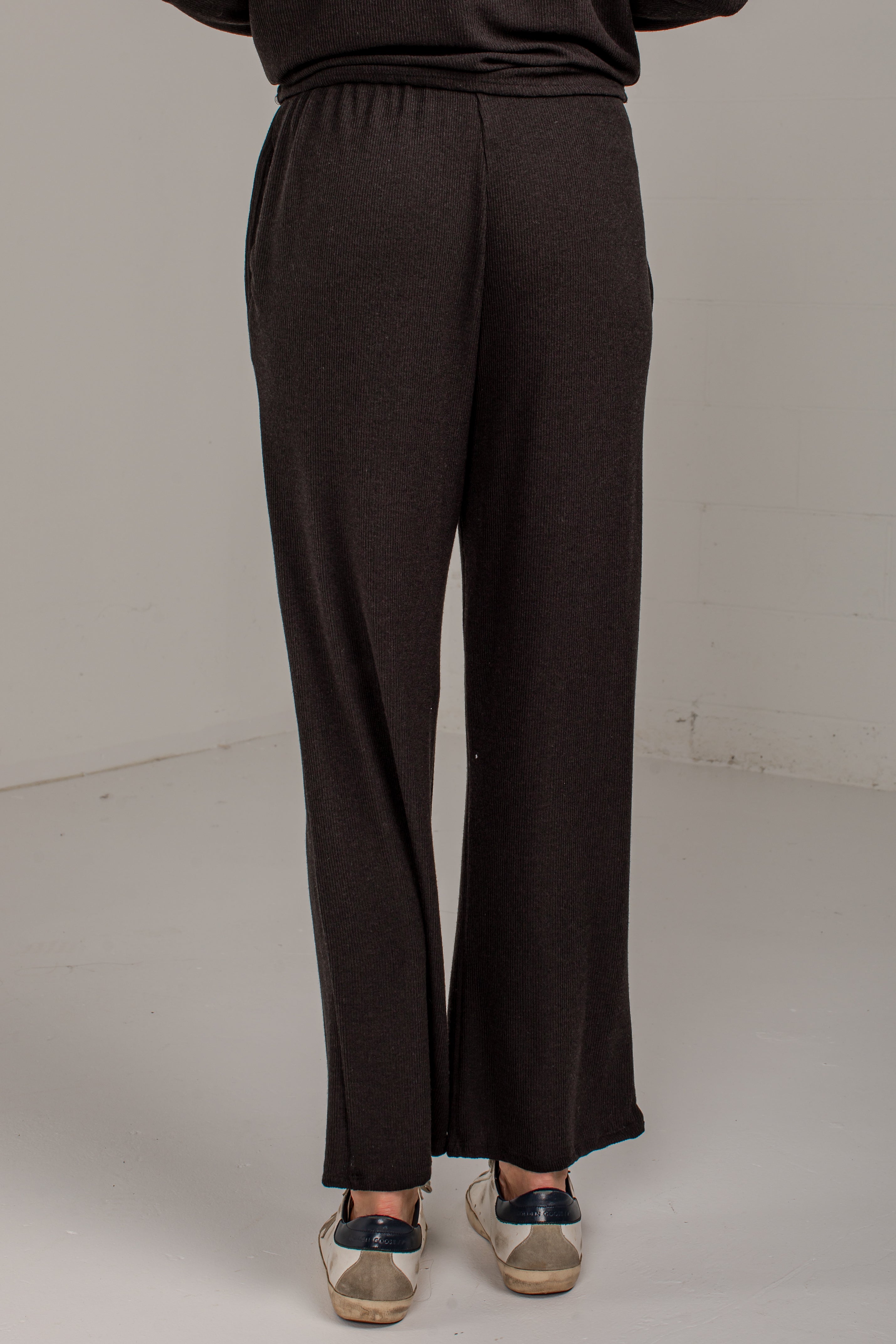 A person shown from the back wearing dark brown wide-leg knit pants and beige sneakers with navy accents, standing against a plain light gray studio background.