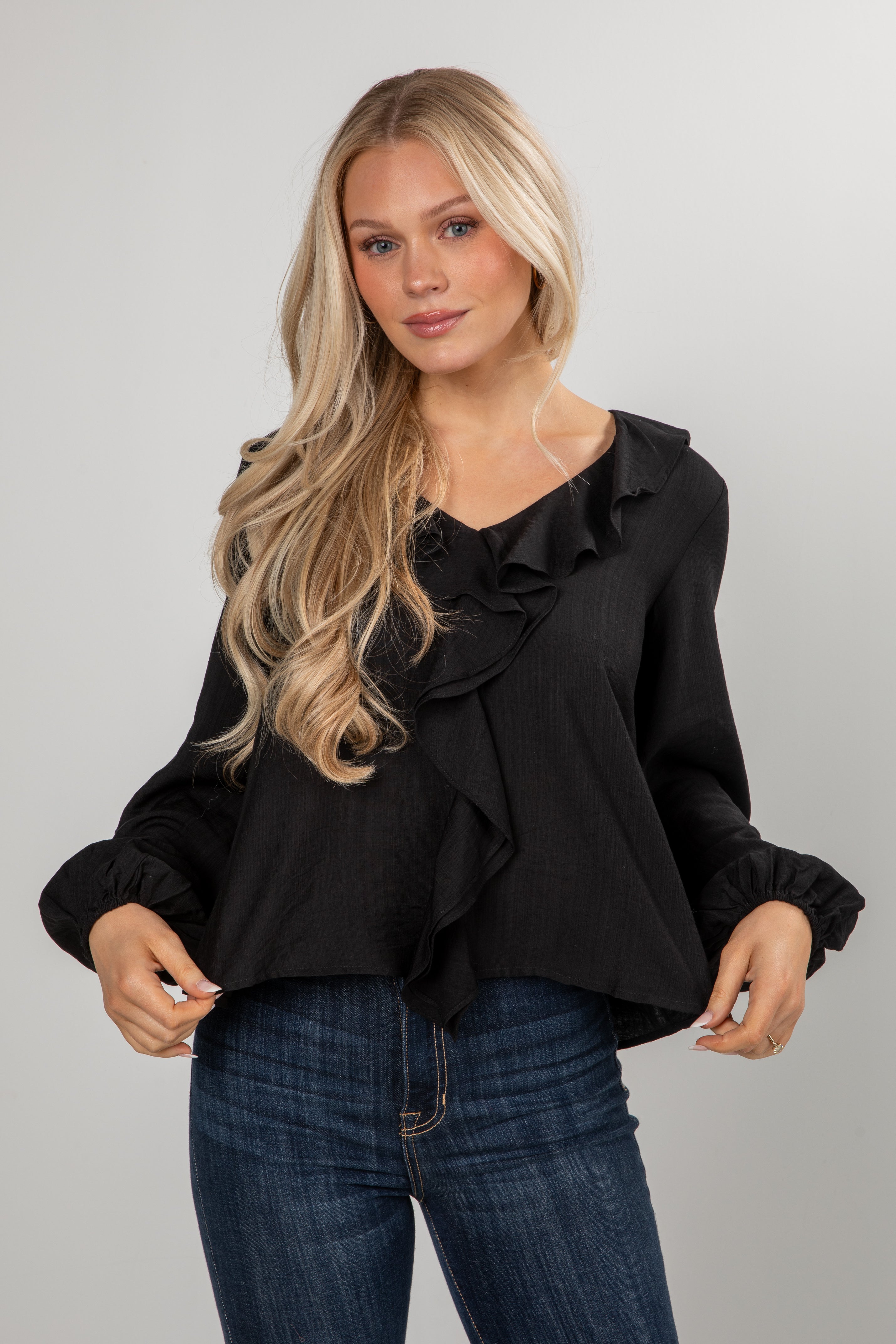 Woman wearing a black blouse with ruffle details and blue jeans on a gray background
