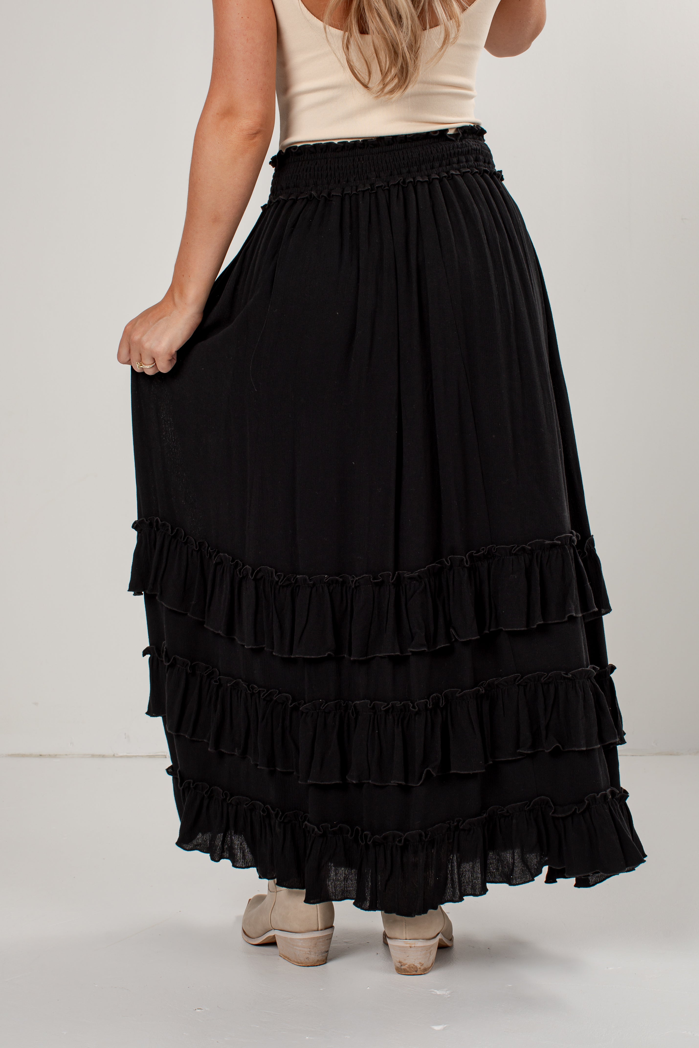 A black tiered maxi skirt with a smocked waistband, shown on a model from the back against a light gray studio background.