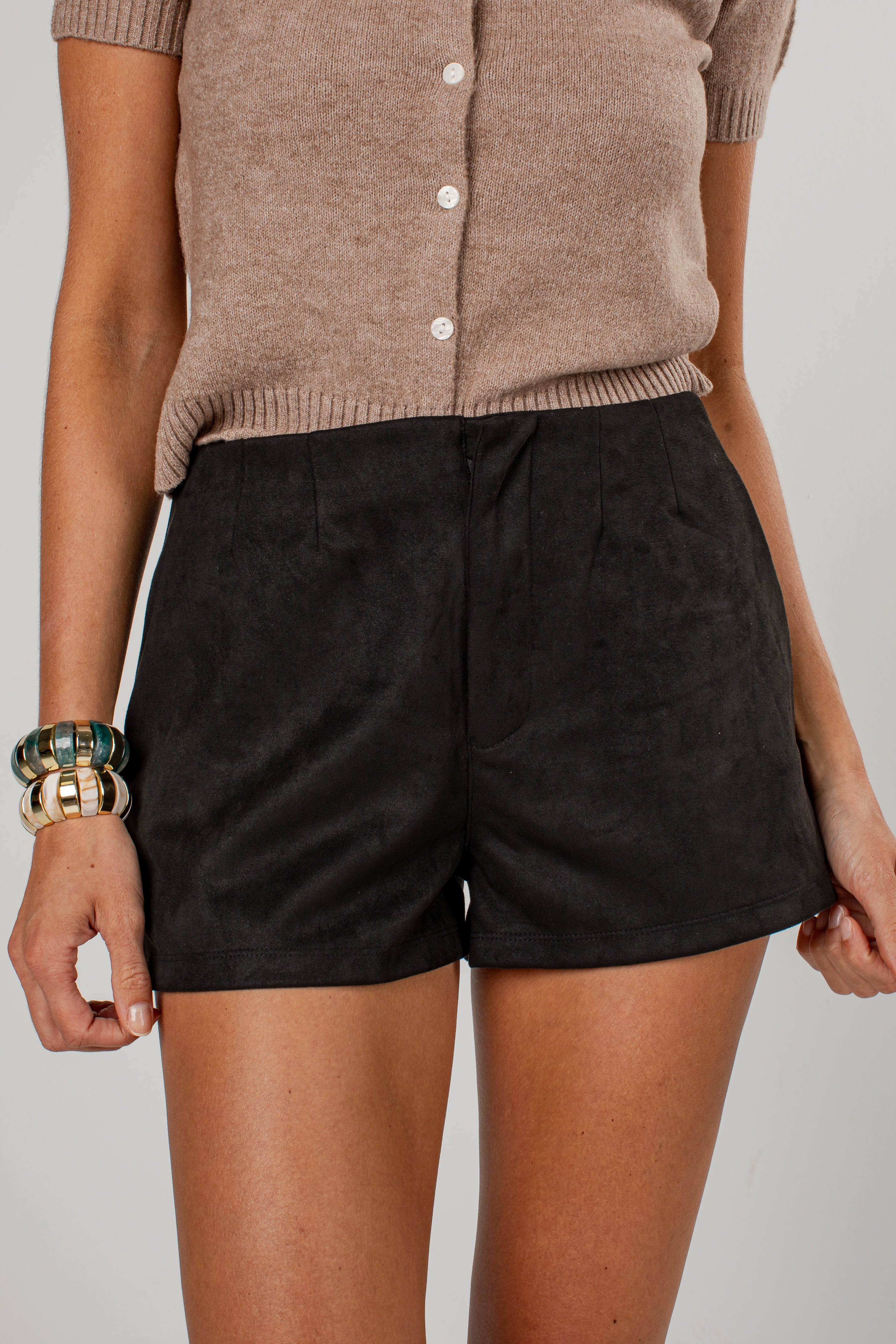 A person wearing black high-waisted shorts and a beige short-sleeve button-front top, shown from the chest to mid-thigh against a plain light gray studio background.