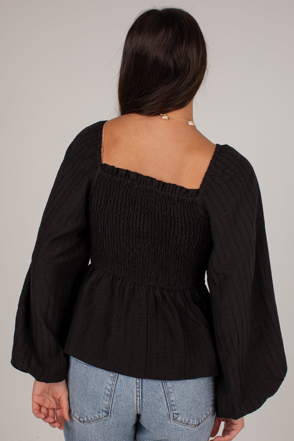 A woman shown from the back wearing a black smocked long-sleeve top with a square neckline and flared sleeves, paired with light blue jeans, against a plain light gray background.