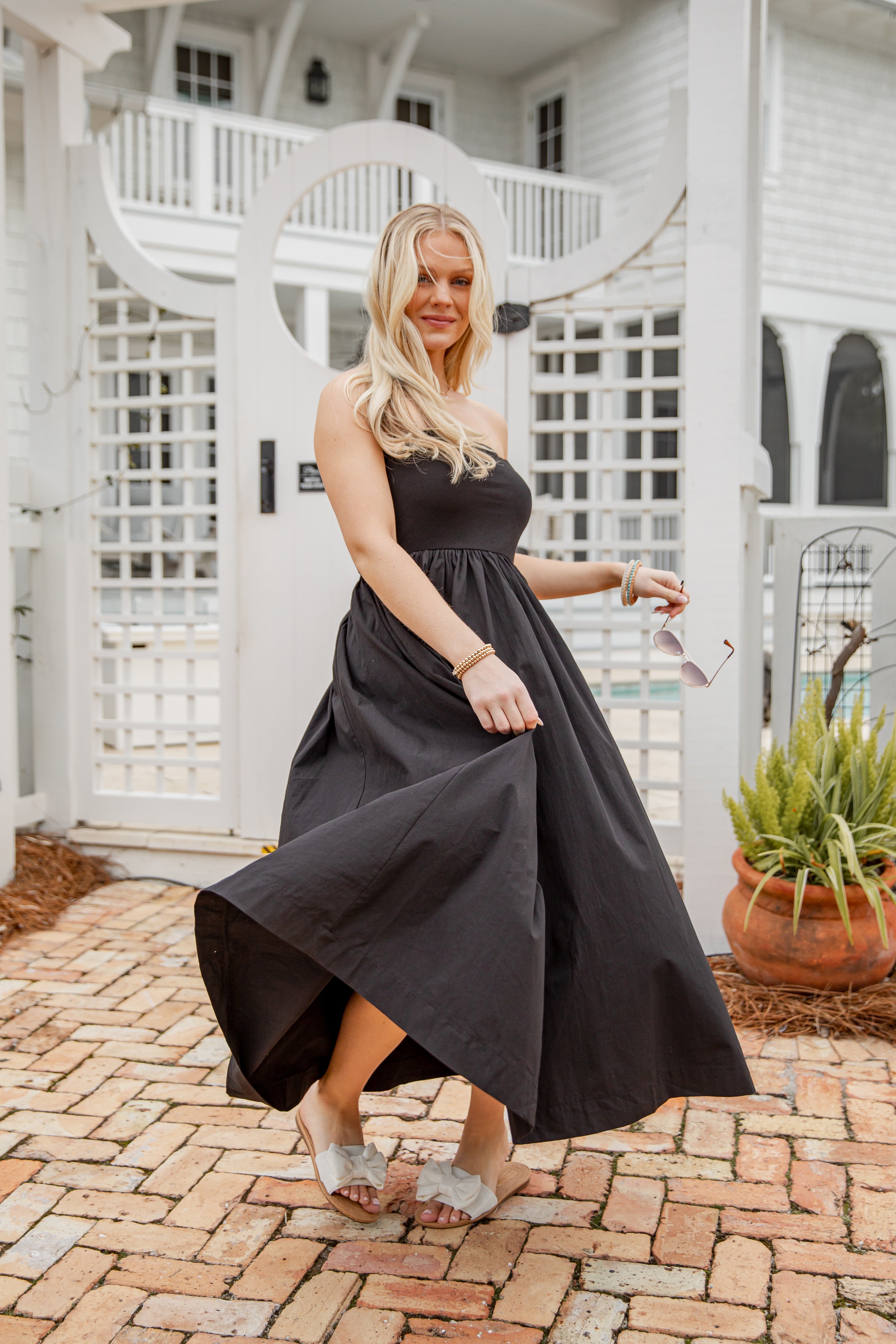 Woman in a black strapless dress standing on a brick patio with a white lattice background.