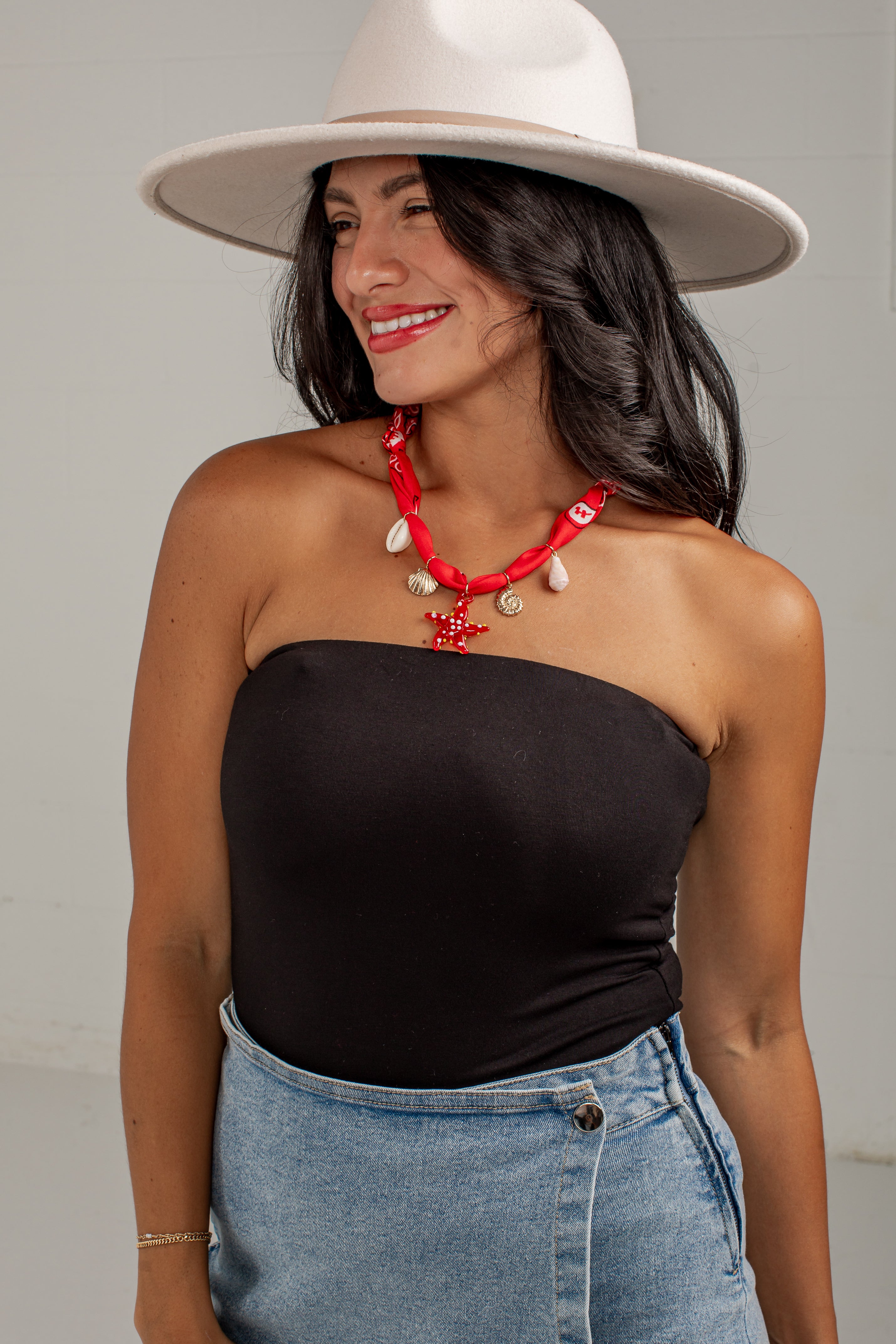 A woman wearing a black strapless bandeau top, light blue denim skirt, white wide-brim hat, and a red beaded necklace with white shell charms, posing against a light gray wall.