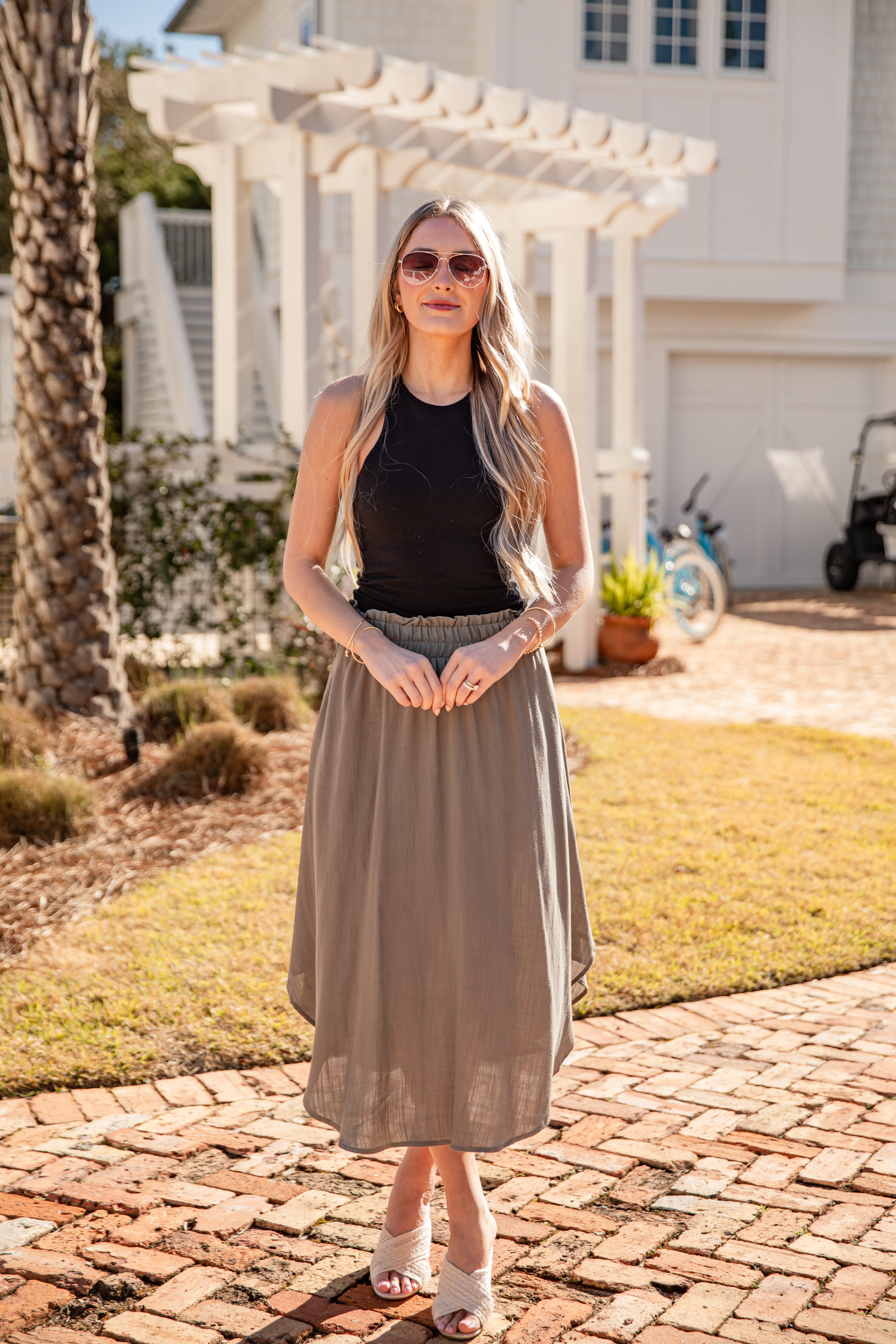Woman standing on a brick path wearing a black top and beige skirt with sunglasses.