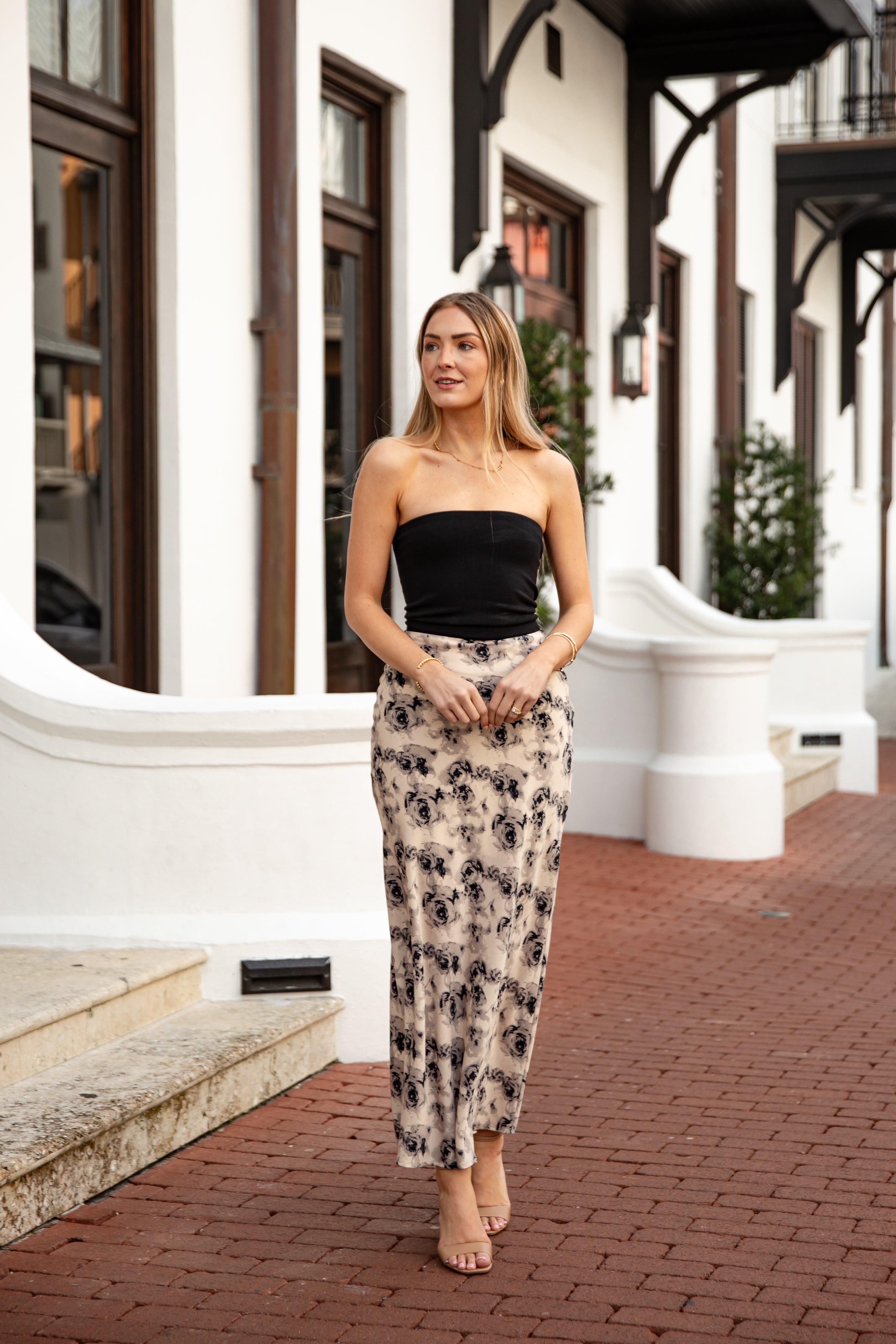 Woman wearing a black strapless top and floral skirt standing on a brick sidewalk.