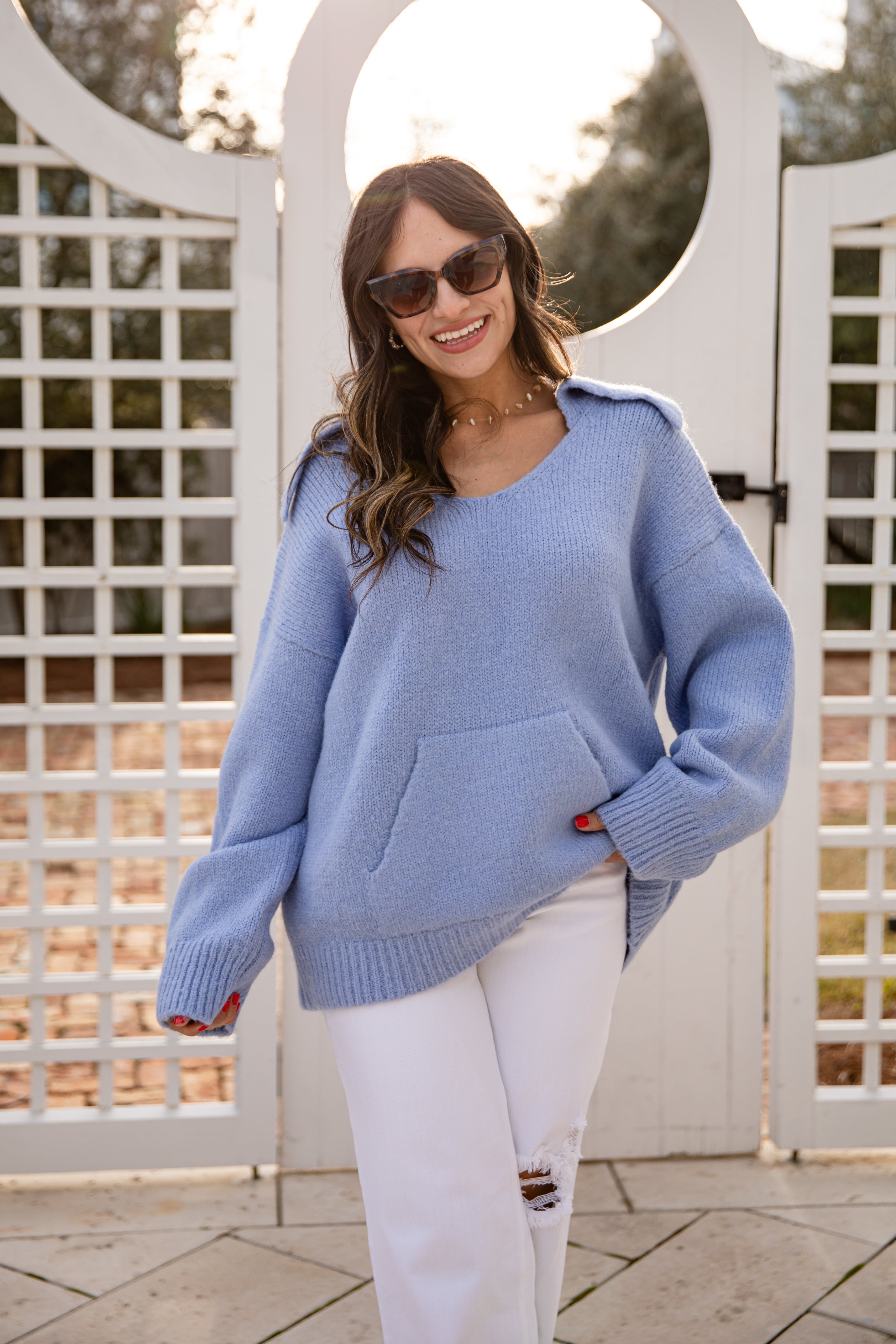 Woman wearing a light blue sweater and white pants standing in front of a decorative white archway.