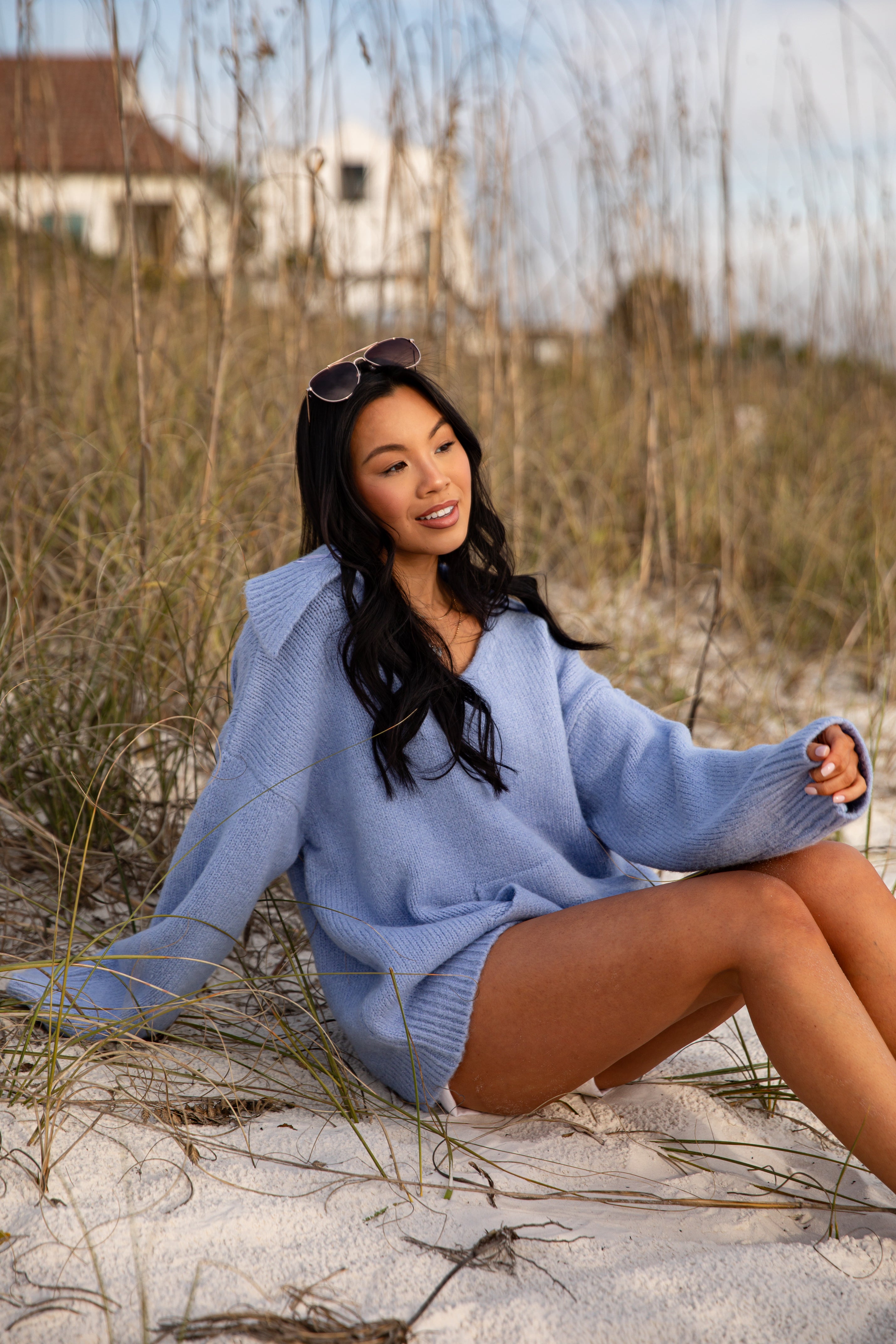 Woman in a light blue sweater sitting on sand with grass and a house in the background