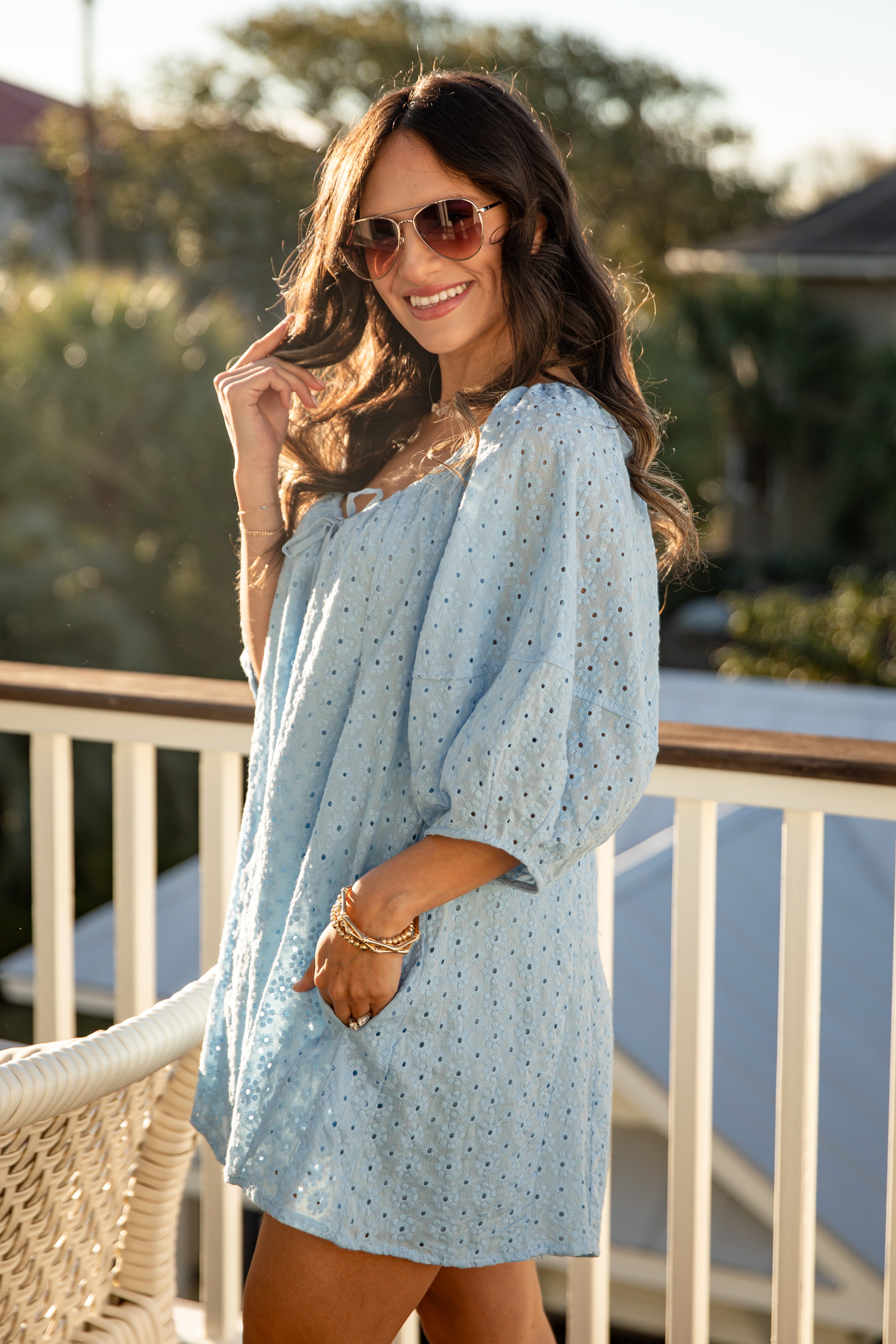 Woman wearing a light blue dress with a pattern, standing on a balcony with greenery in the background.