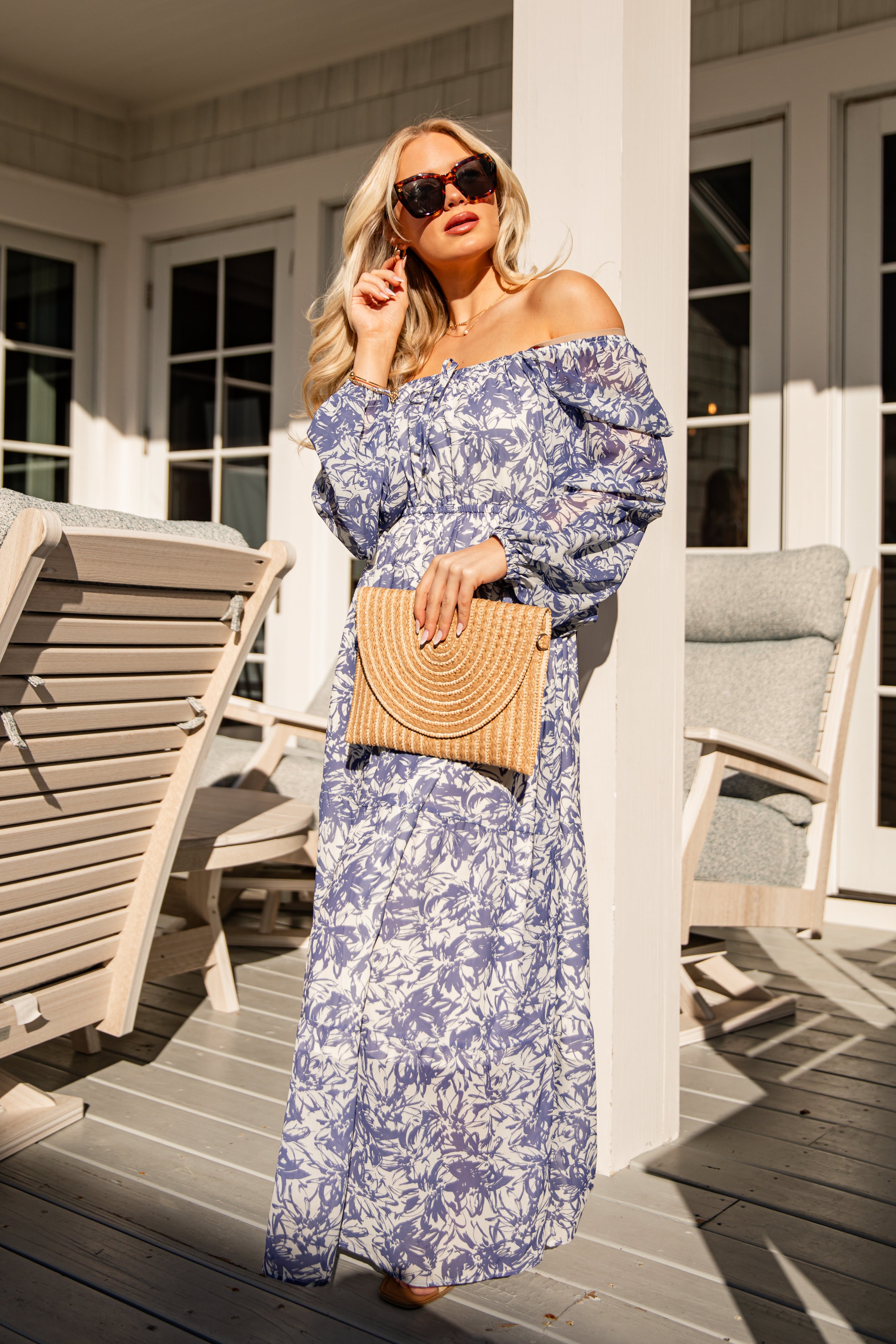 Woman in a blue floral dress holding a woven handbag on a porch.