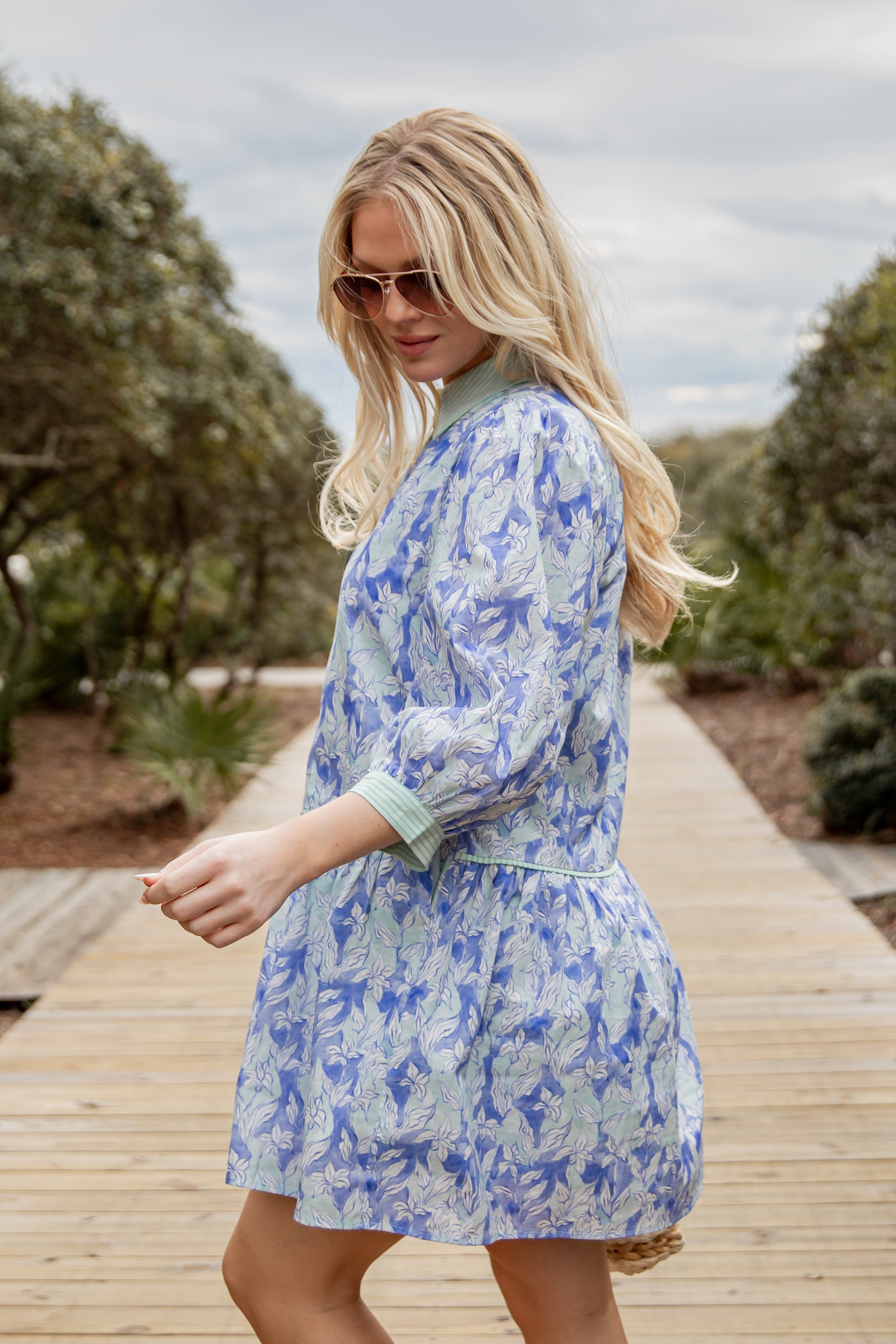 Woman wearing a blue floral dress standing on a wooden boardwalk with trees in the background