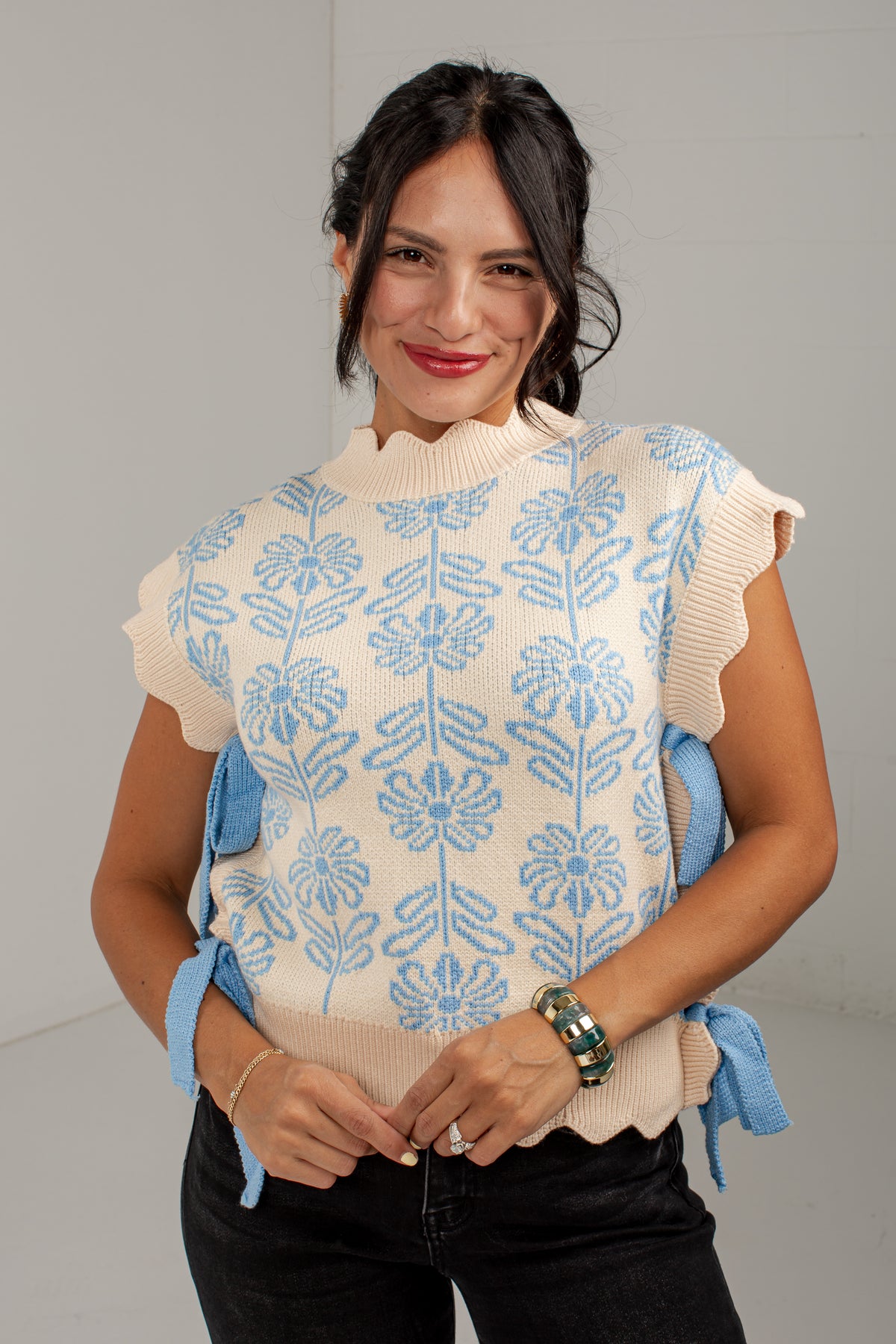 A woman wearing a cream-colored knit sweater vest with a light blue floral pattern and blue tie details at the sides, paired with black jeans.