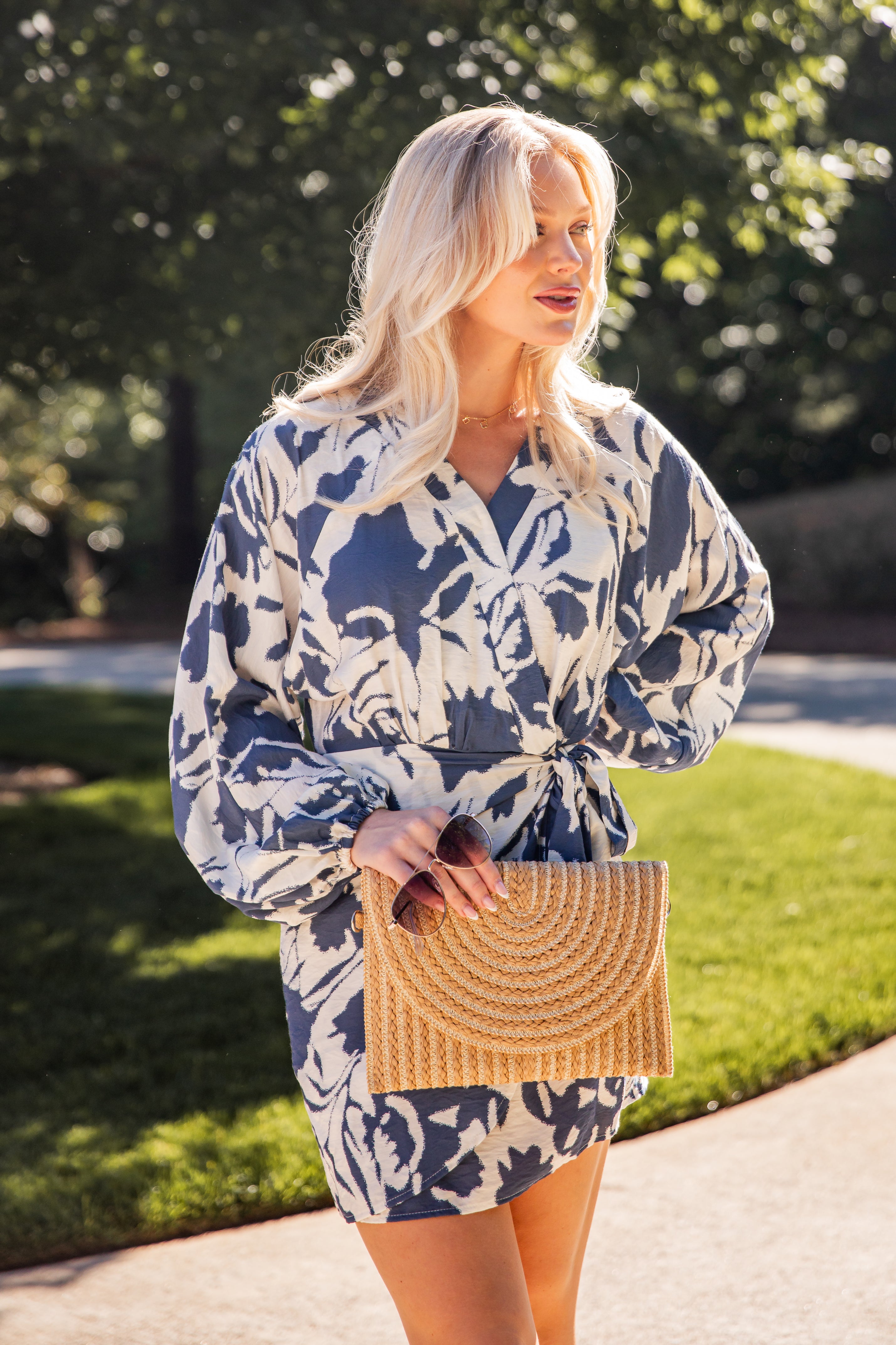 Model wearing a blue floral print wrap mini dress with long sleeves and a waist tie, styled with a woven clutch outdoors.