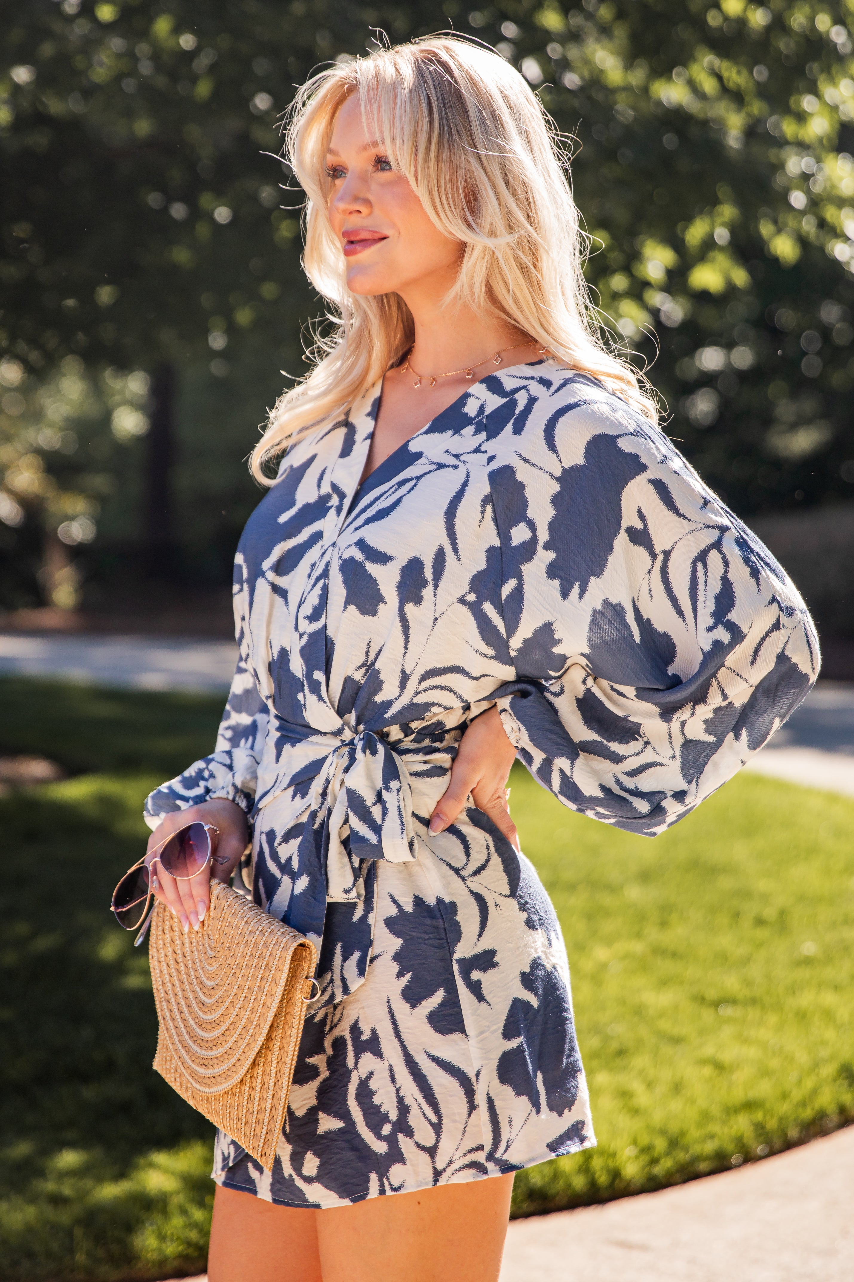Model wearing a blue floral print wrap mini dress with long sleeves and a waist tie, styled with a woven clutch outdoors.