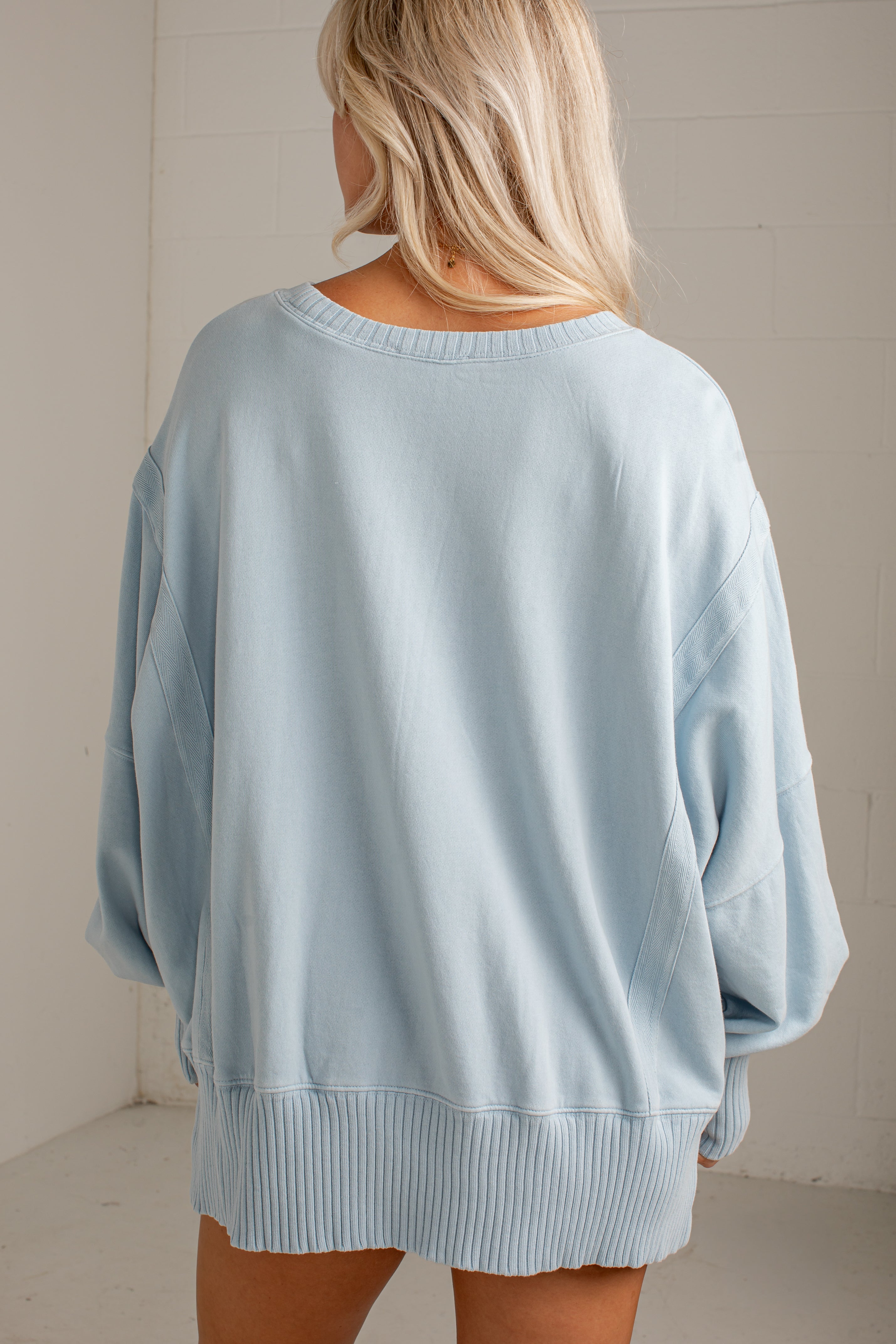 A woman viewed from behind wearing an oversized light blue knit sweater with wide sleeves and a ribbed hem, set against a plain off-white wall.
