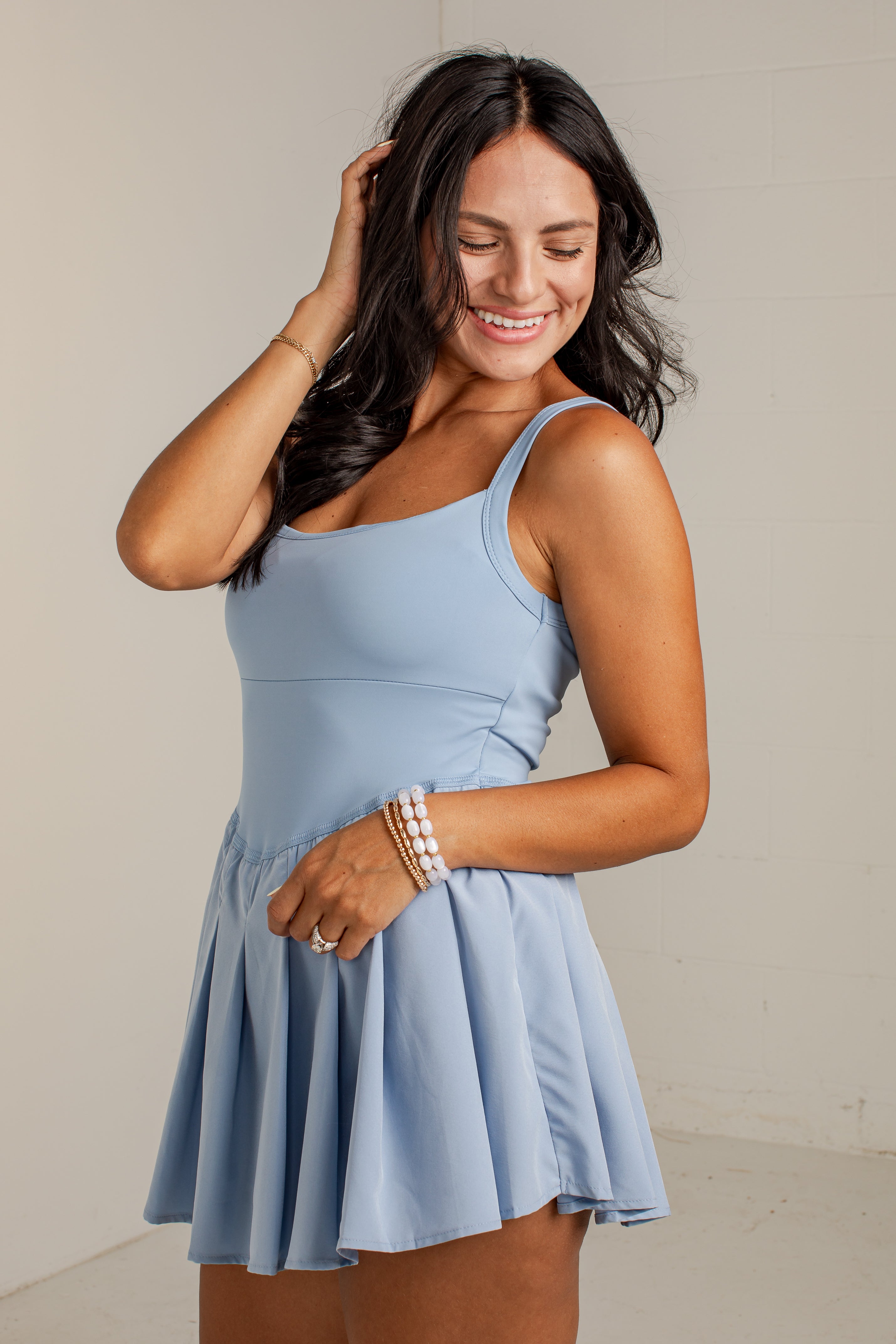 A woman modeling a light blue sleeveless mini dress with a scoop neckline and a flared skirt, standing against a plain off-white wall.