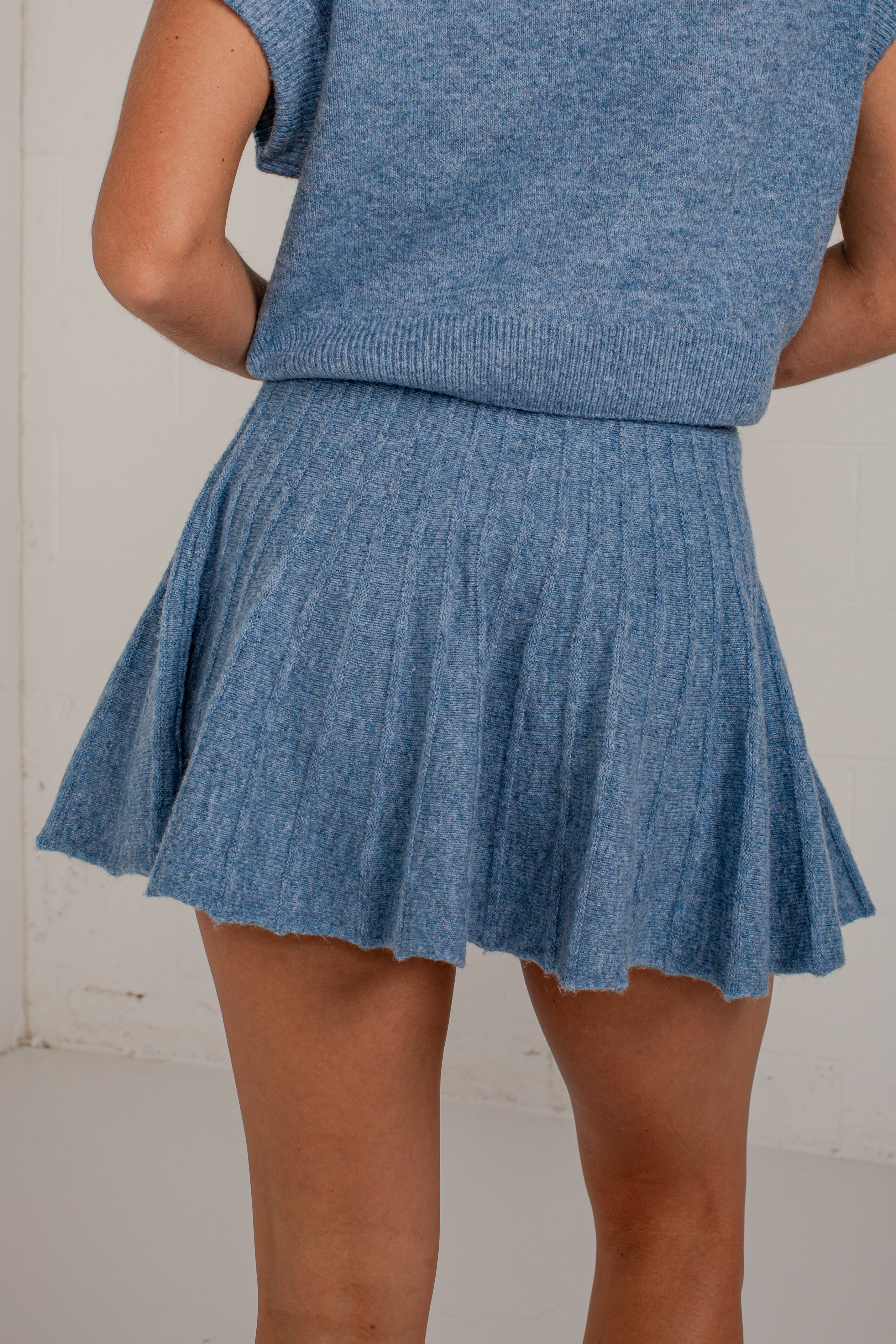 A person wearing a heathered blue ribbed knit sweater vest and matching pleated mini skirt, shown from the back against a light neutral wall.