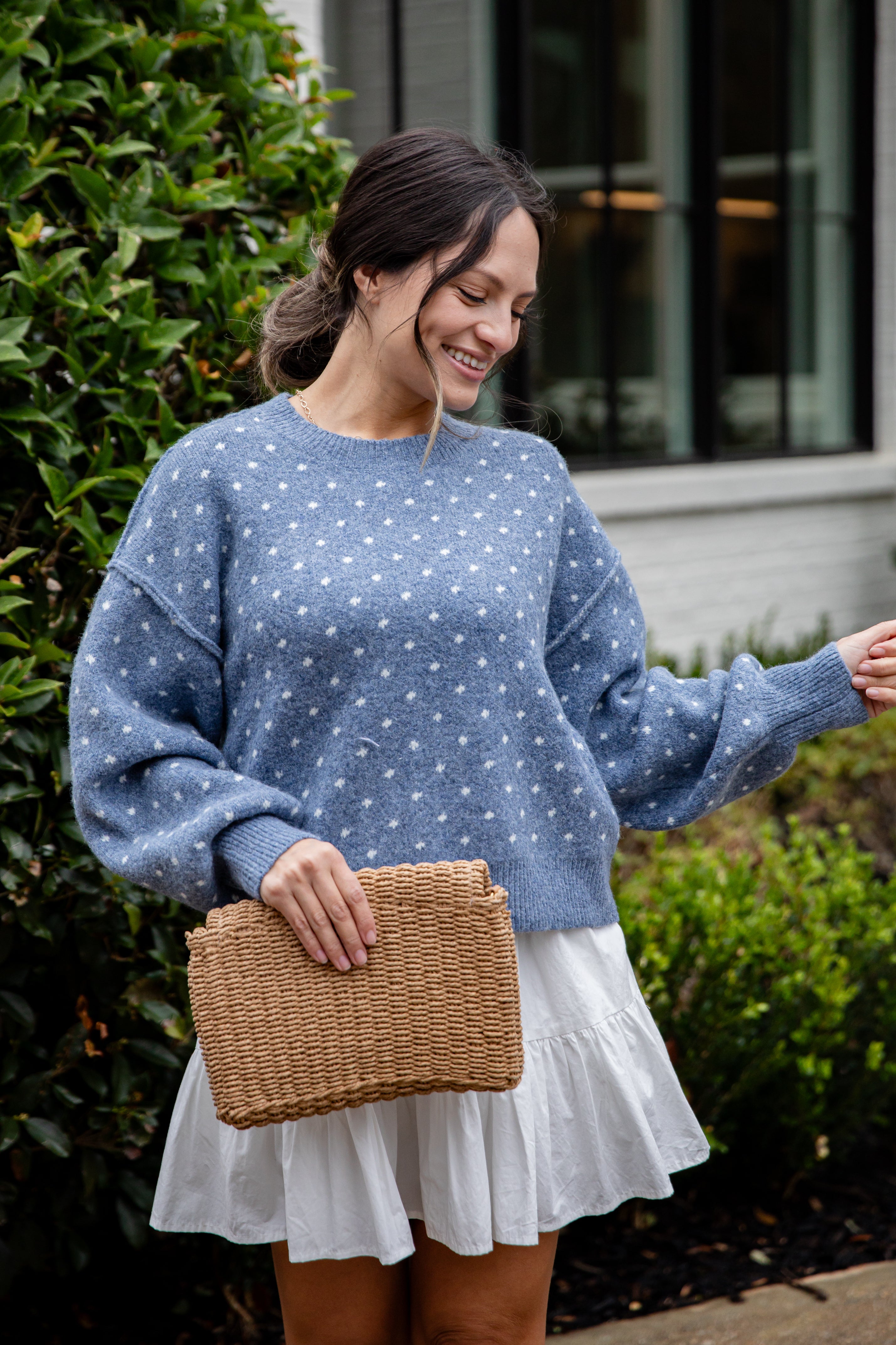 Woman wearing a blue polka dot sweater and white skirt, holding a woven bag outdoors.