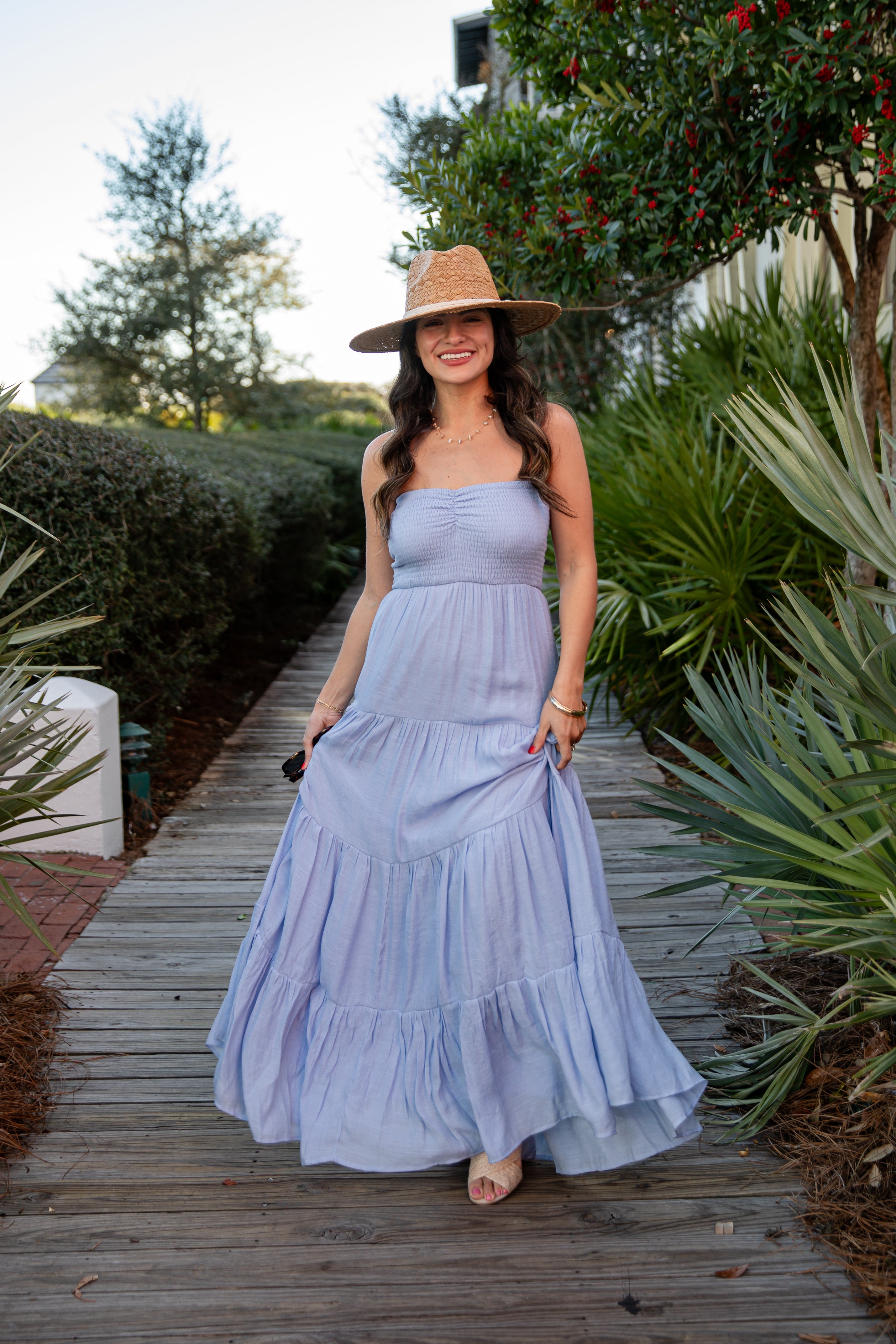 Woman in a light blue dress and hat standing on a wooden path with greenery around.