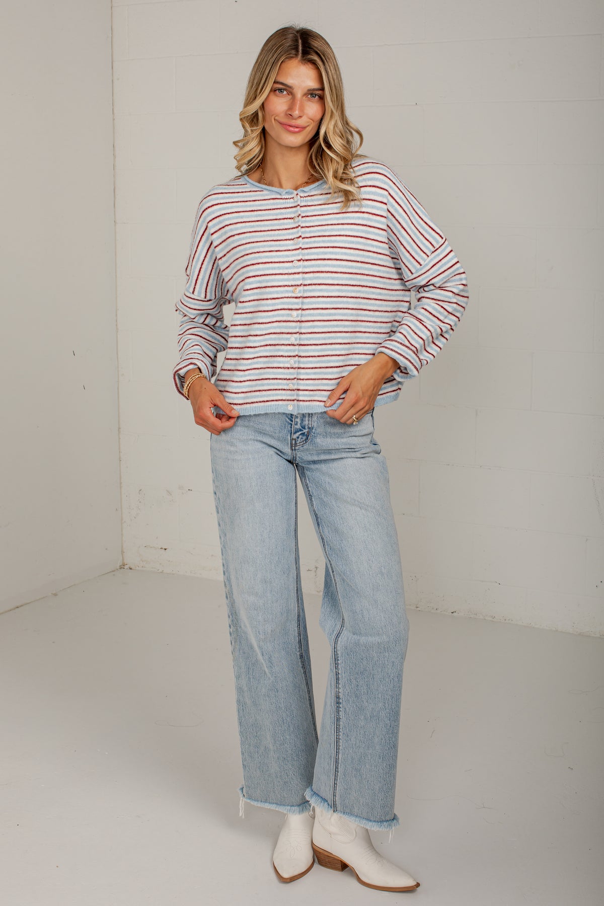 A woman modeling a long-sleeve button-up shirt with thin red and orange horizontal stripes on a white background, paired with light-wash wide-leg jeans and white ankle boots.