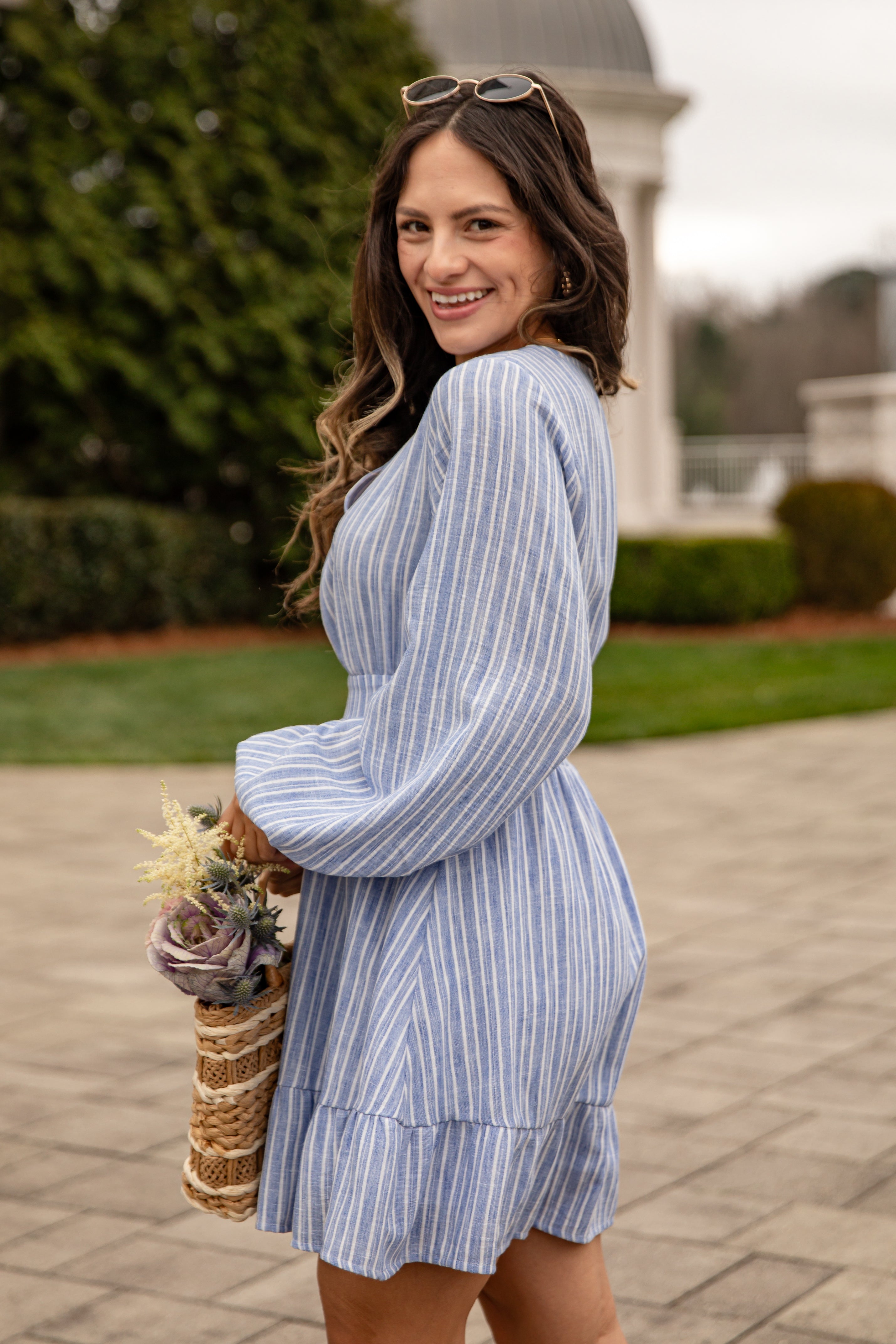 Woman in a blue striped dress holding a woven basket with flowers outdoors.