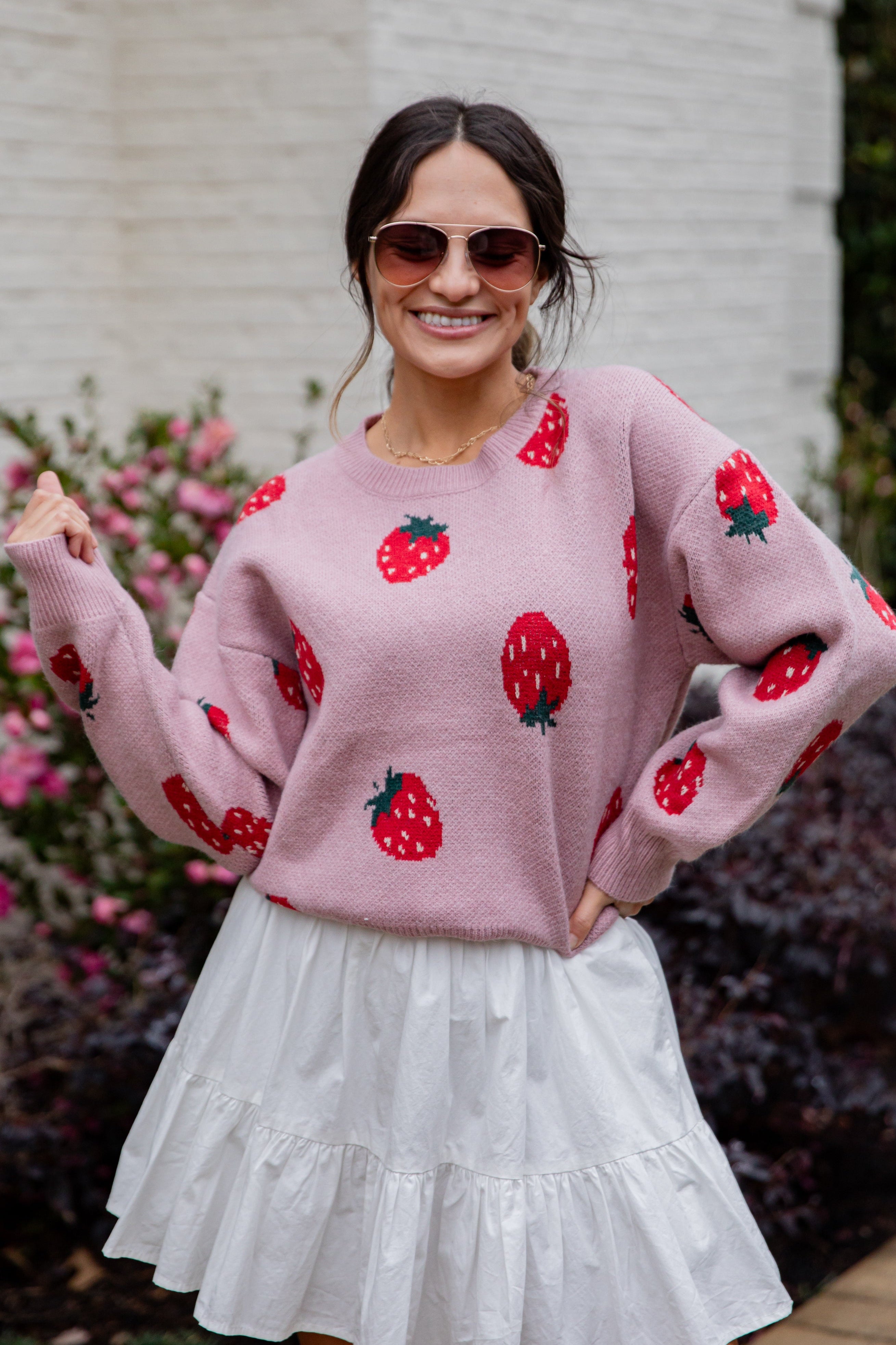Woman wearing a pink sweater with strawberry patterns and a white skirt outdoors.
