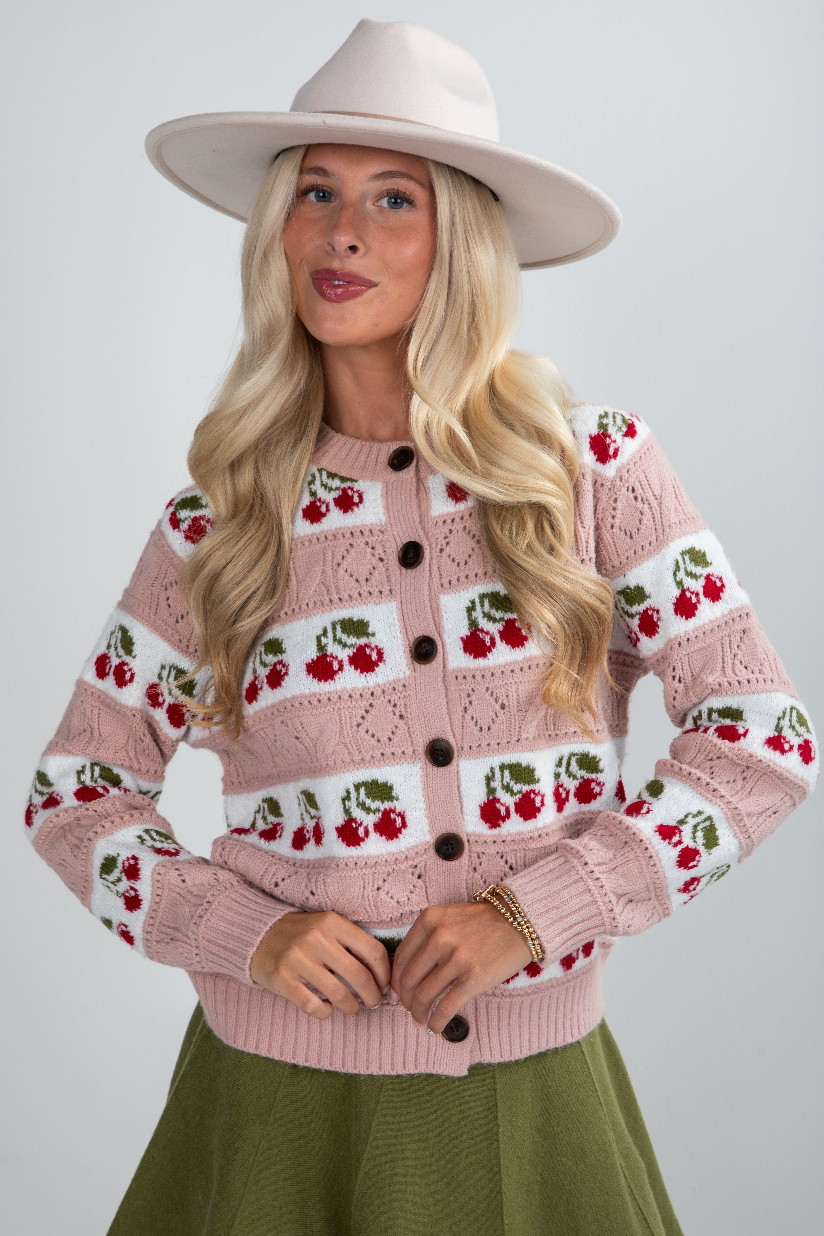 Woman wearing a pink knitted cardigan with cherry pattern, beige hat, and green skirt on a white background