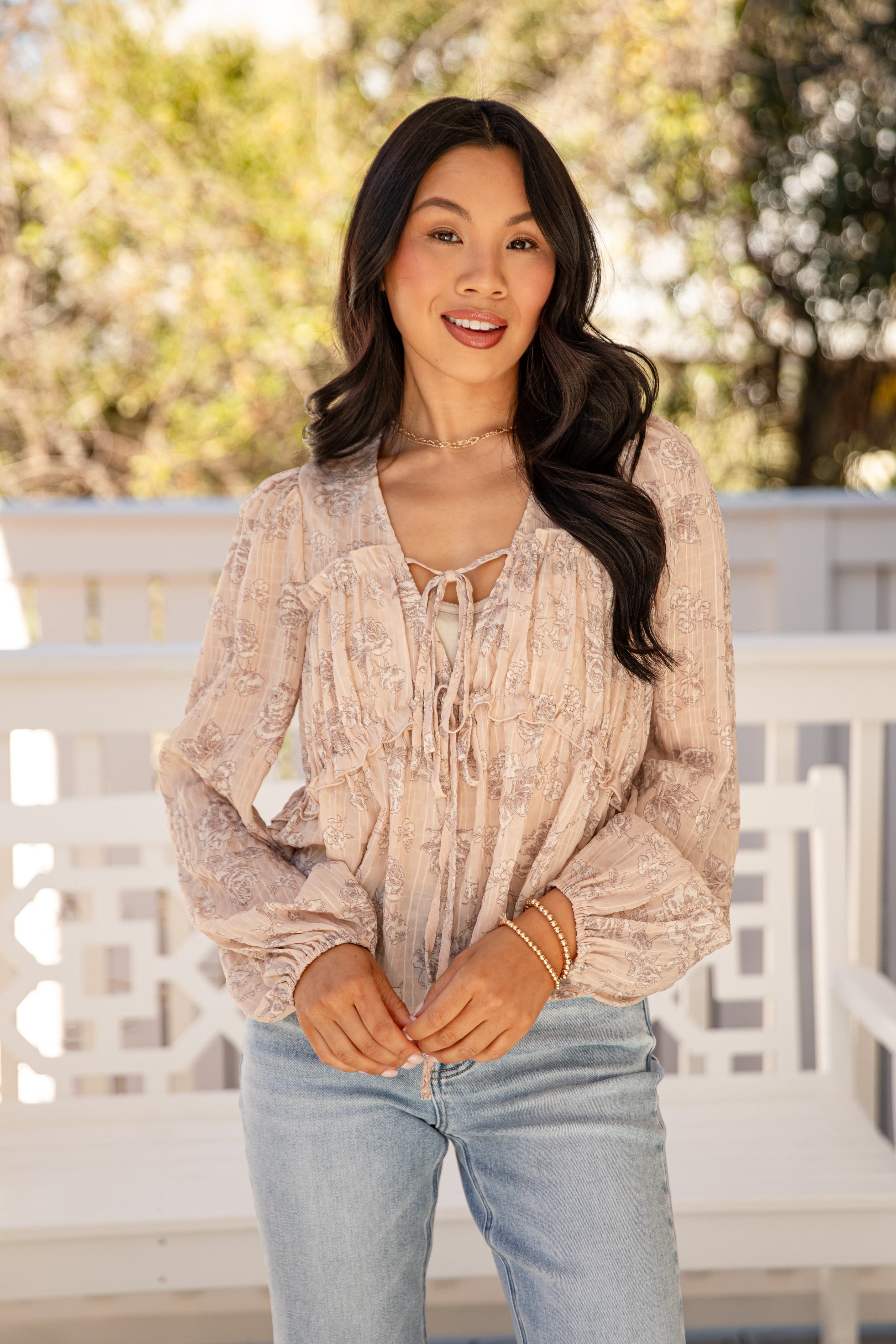 Woman wearing a beige blouse and light blue jeans standing outdoors with blurred greenery in the background