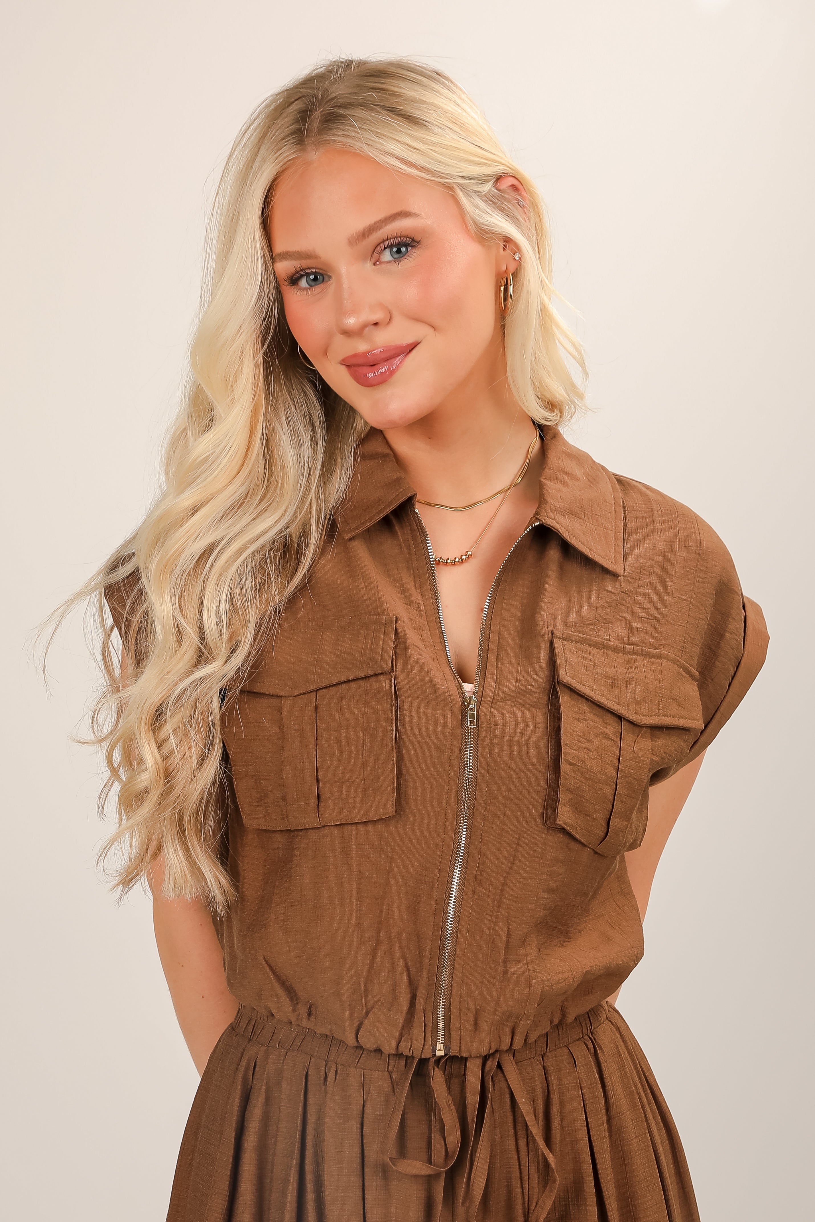Woman wearing a brown sleeveless jacket over a dress against a plain background