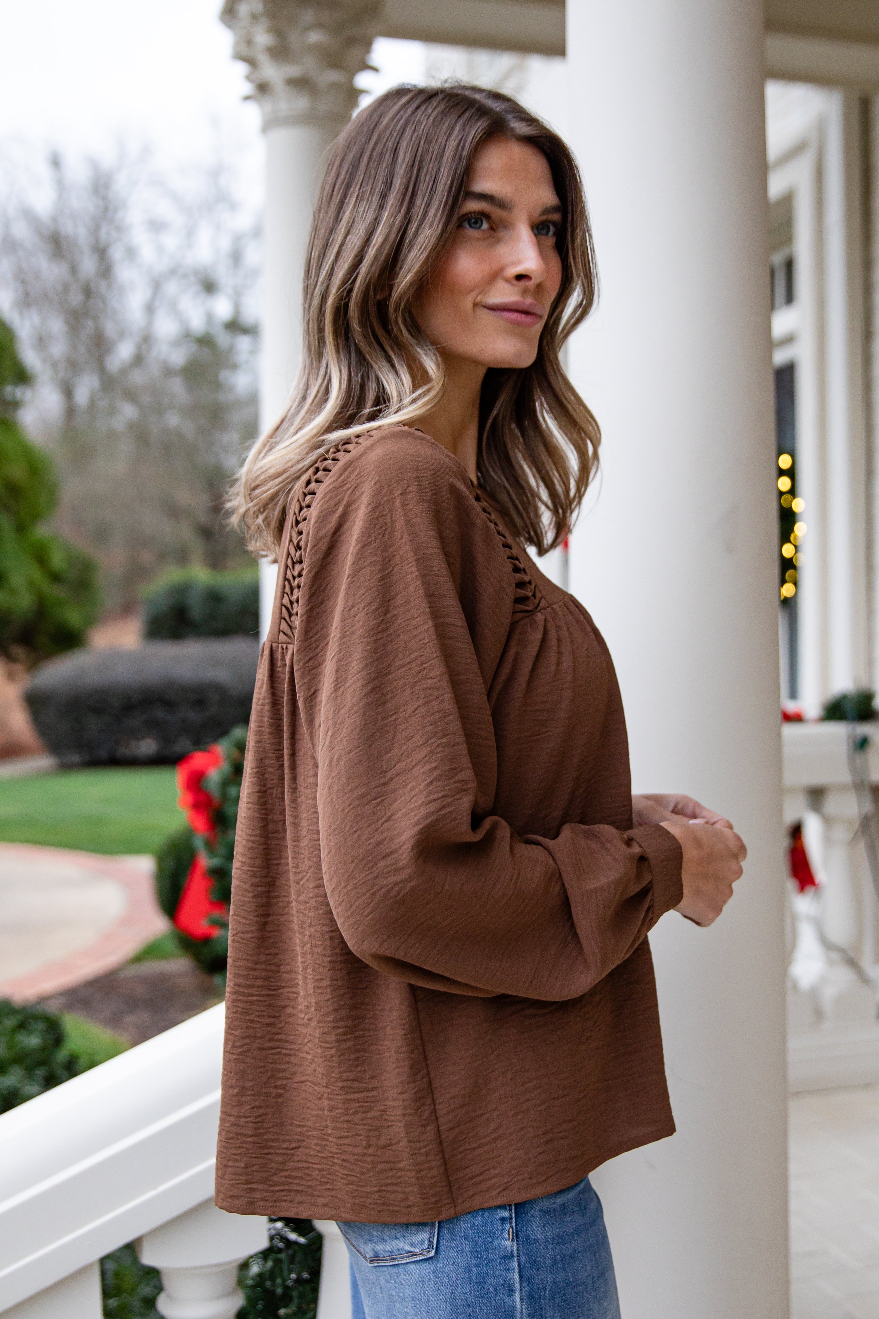 Woman wearing a brown sweater and blue jeans standing on a porch.