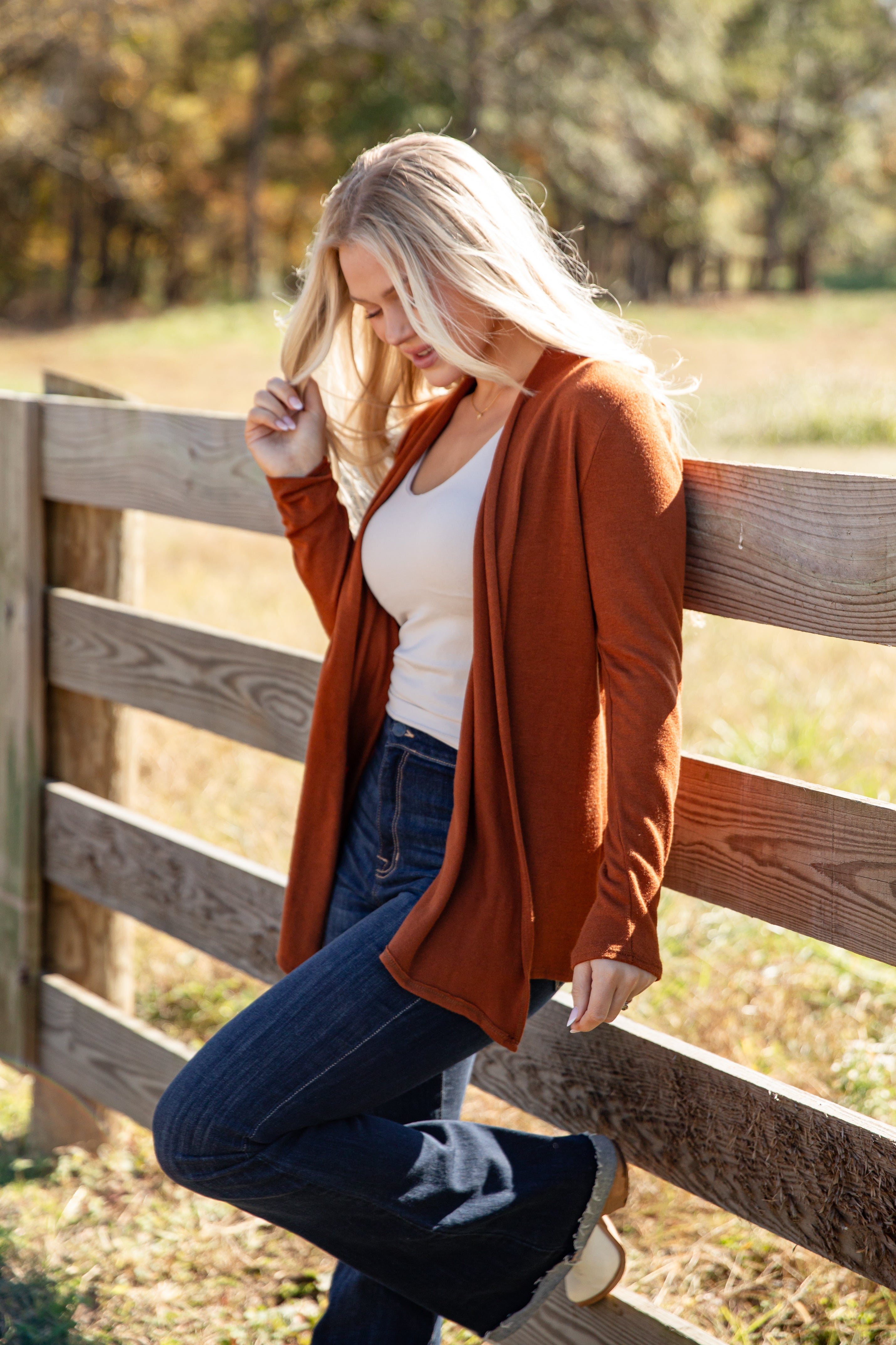 Woman wearing a rust-colored cardigan, white top, and blue jeans standing by a wooden fence.