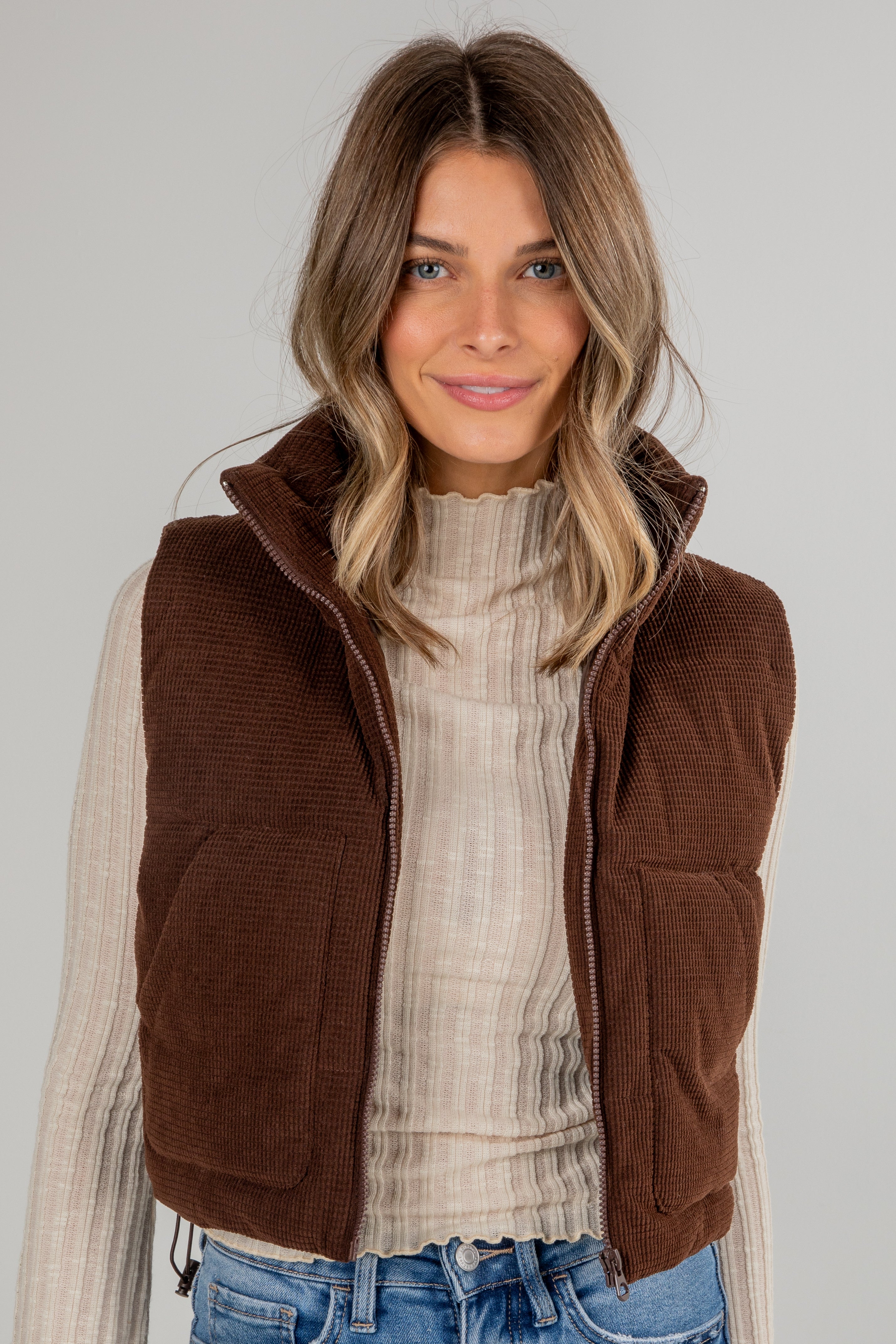 Woman wearing a brown vest over a white sweater and blue jeans on a plain background