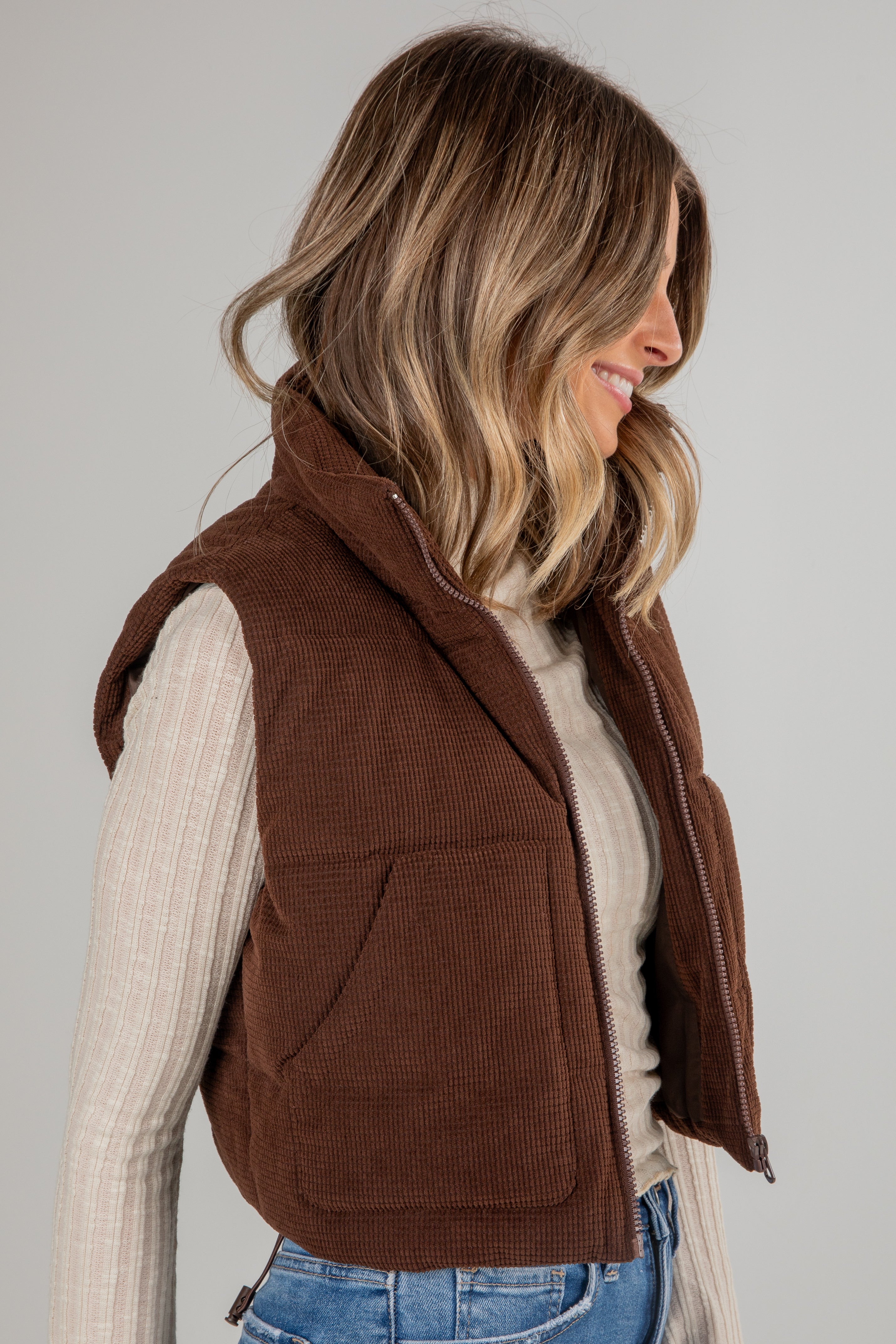 Woman wearing a brown vest over a beige sweater and blue jeans on a plain background