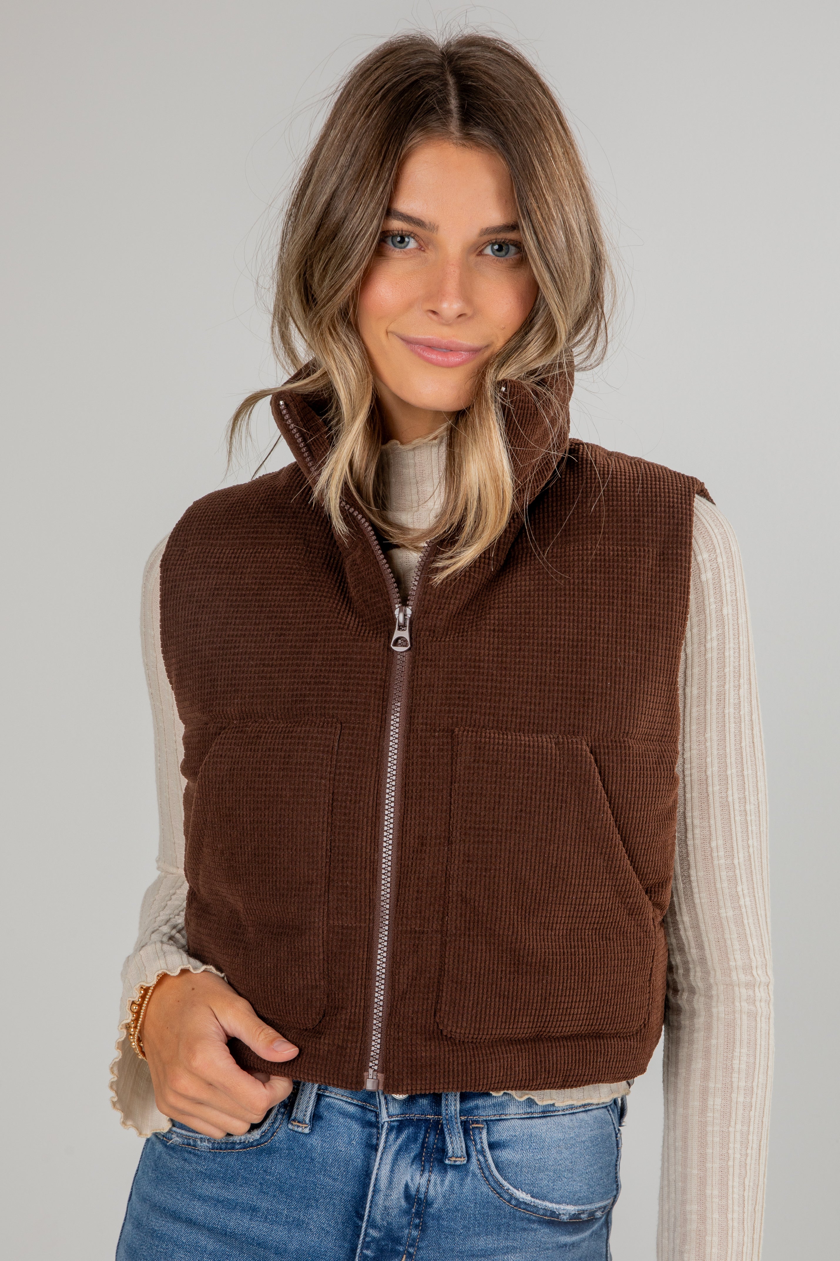 Woman wearing a brown corduroy vest over a white shirt and blue jeans on a plain background