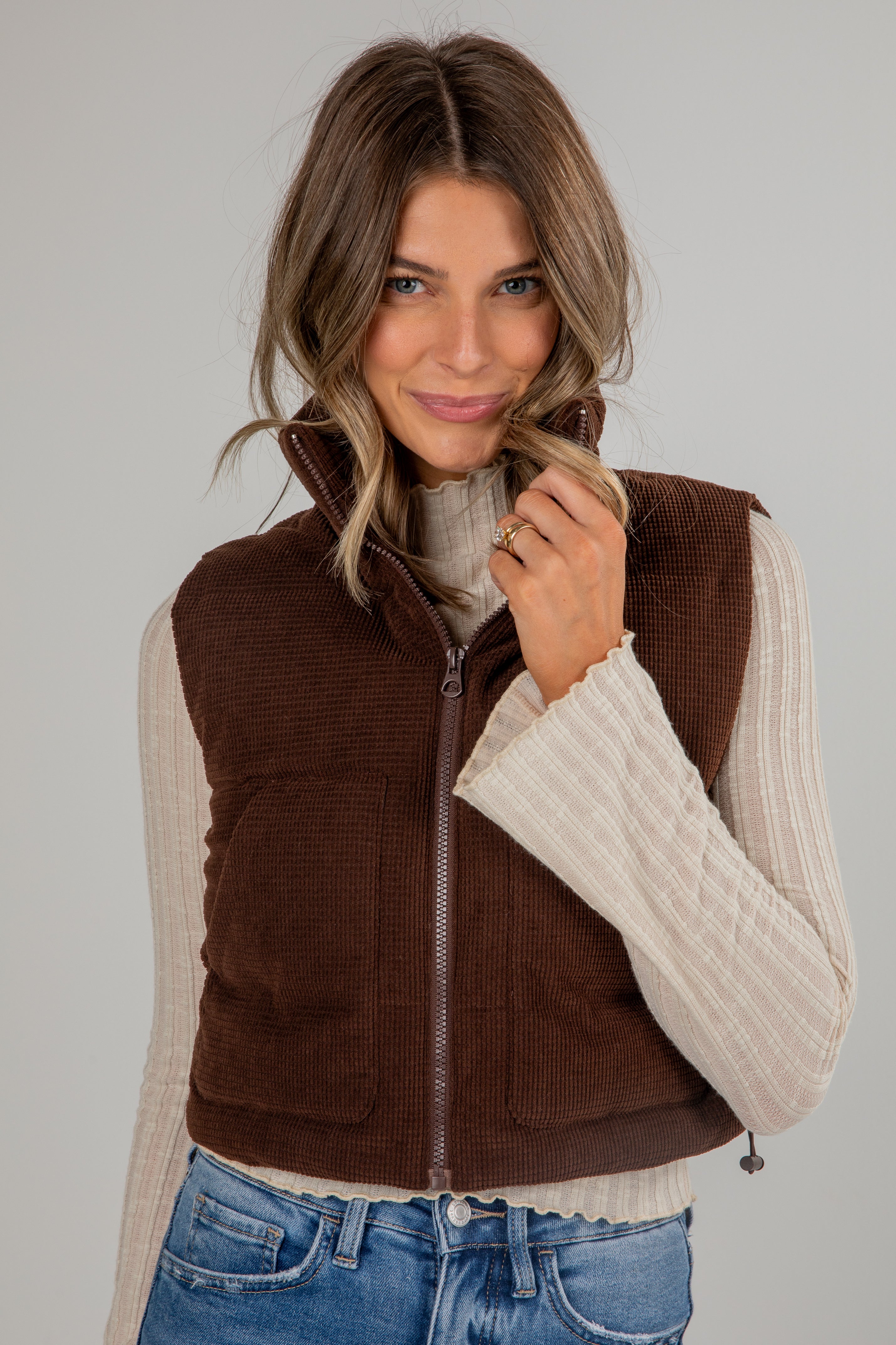Woman wearing a brown vest over a cream sweater with a plain background