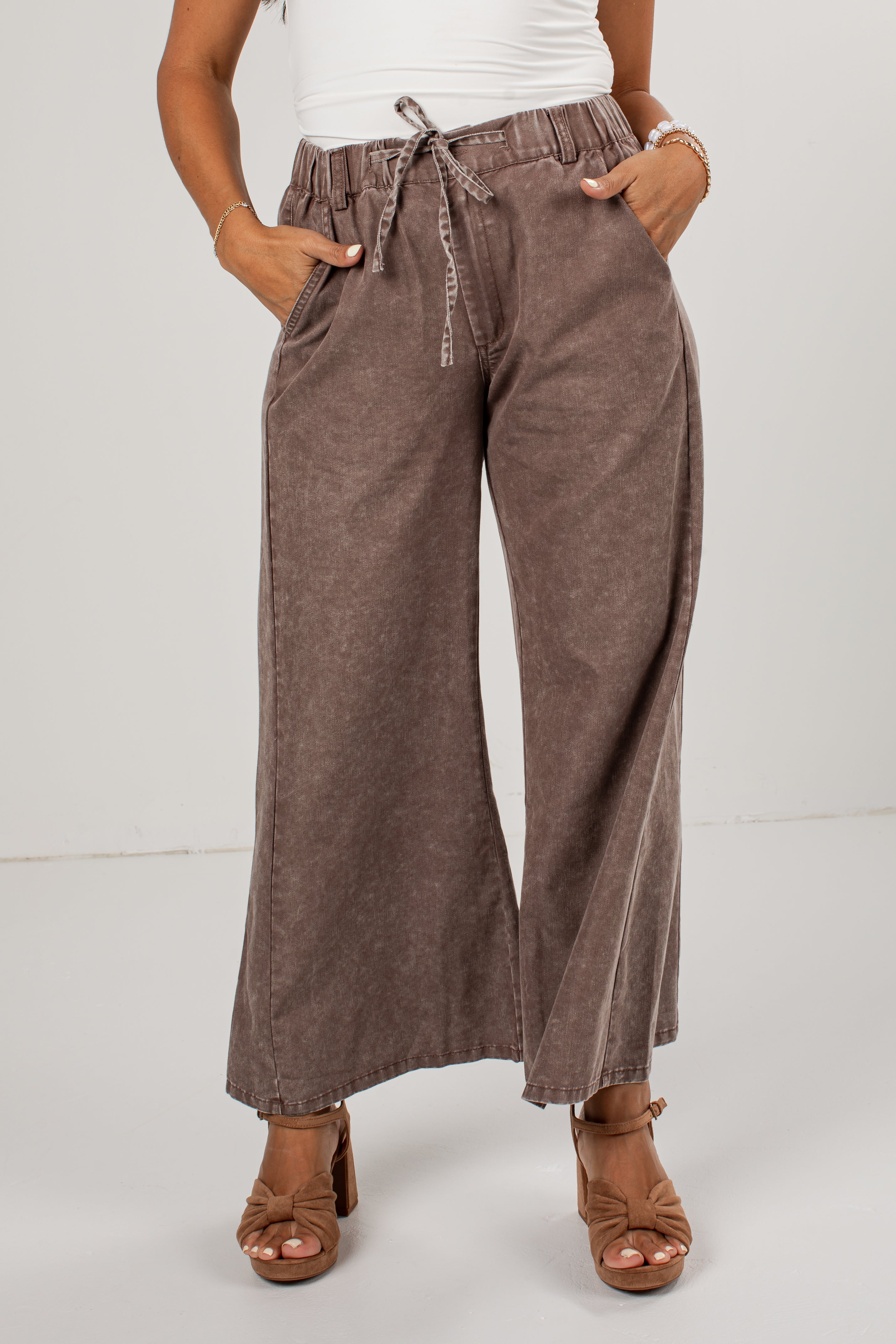 A person wearing high-waisted, wide-leg brown pants with a drawstring waist, paired with a white top and brown heeled sandals, photographed against a plain light gray studio background.