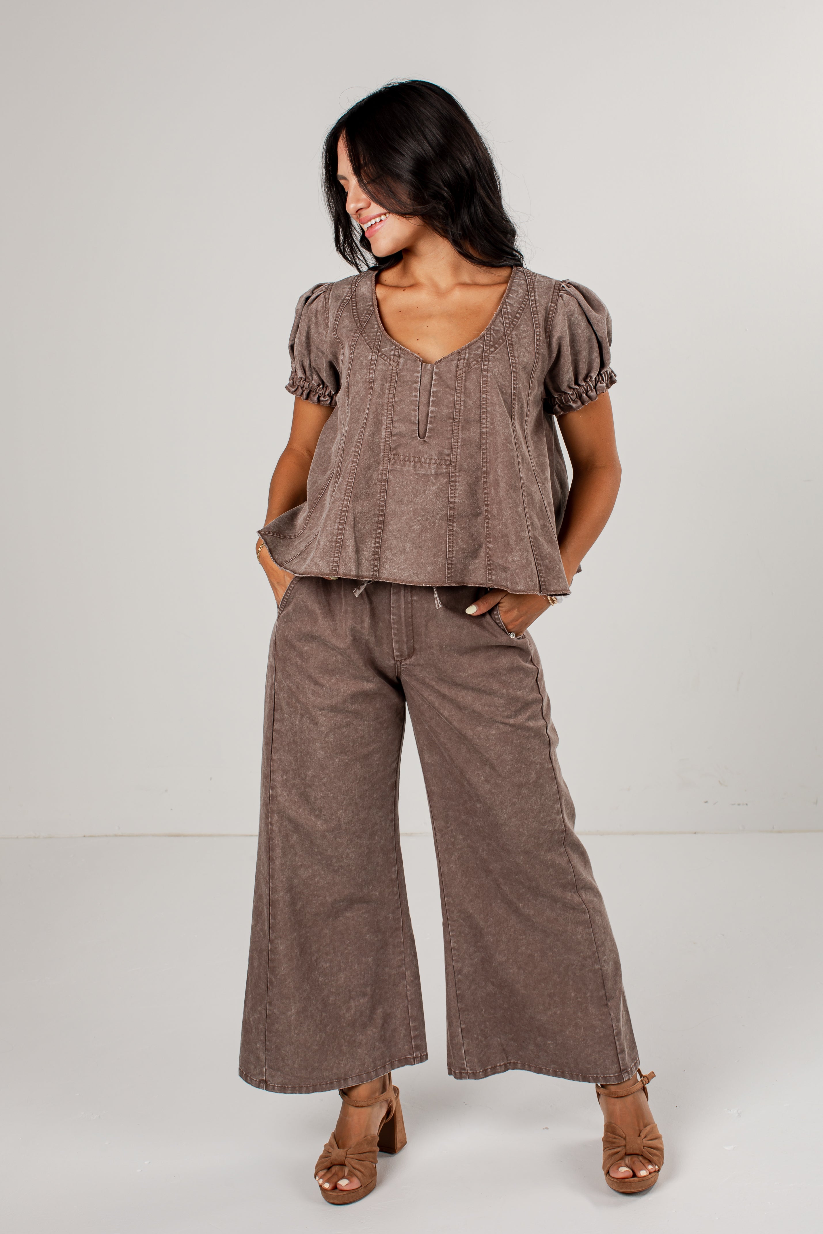 A woman modeling a matching two-piece outfit consisting of a short-sleeve peplum top and wide-leg pants in a textured taupe-brown fabric, paired with brown heeled sandals against a plain light gray studio background.