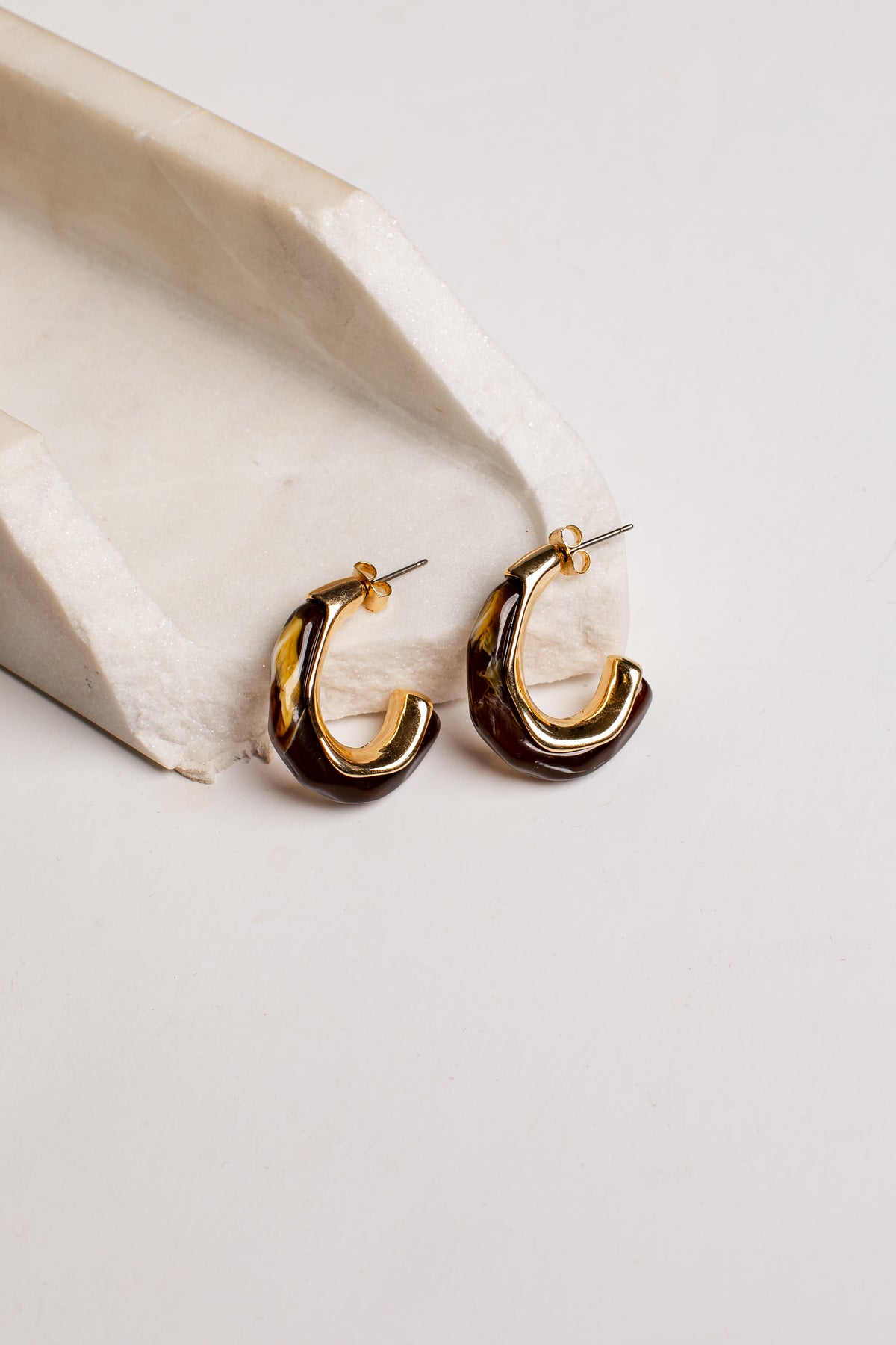 A pair of gold and dark brown tortoiseshell hoop earrings resting on a white textured stone prop against a light gray background.