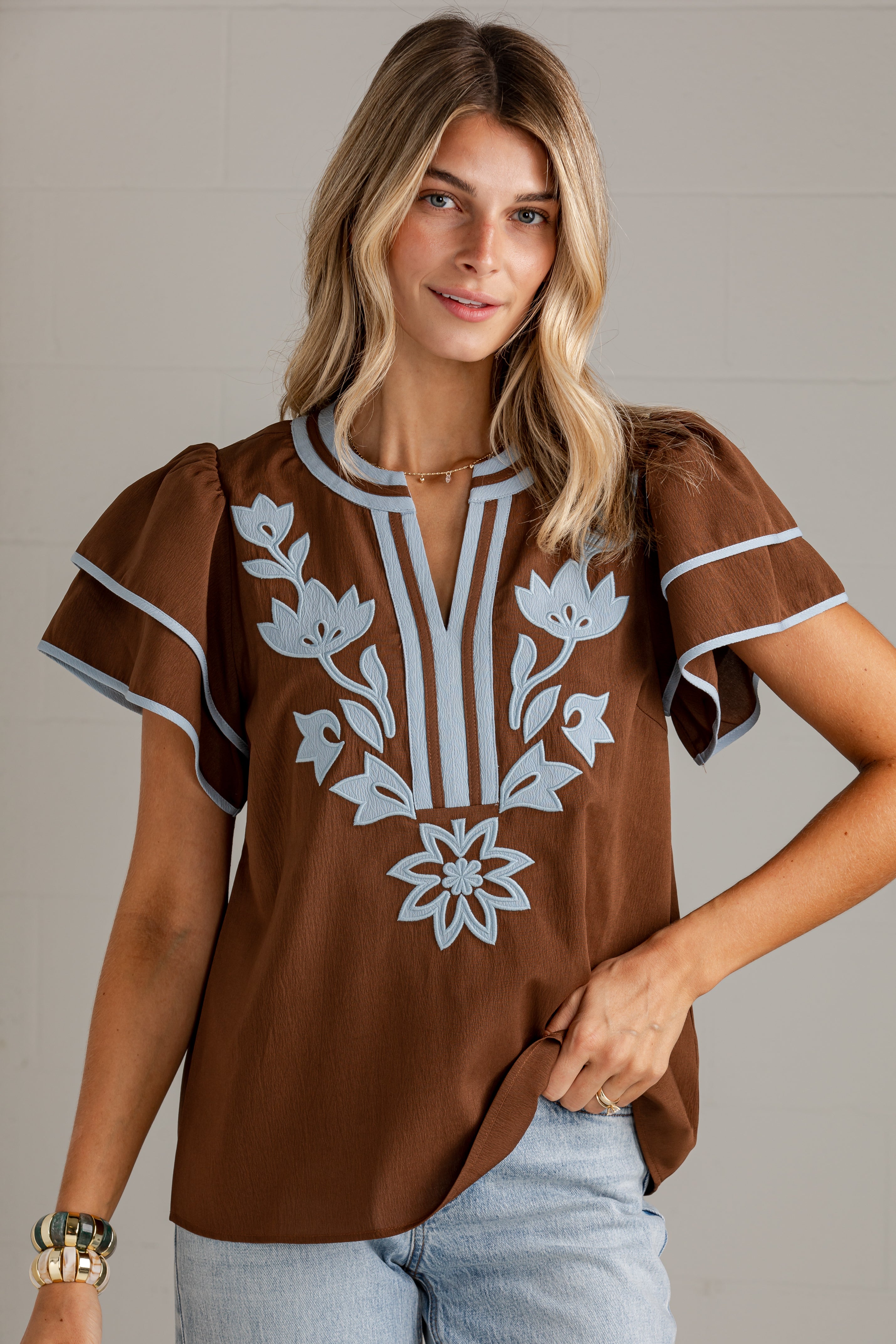A woman wearing a brown short-sleeve blouse with white floral embroidery and striped trim, paired with light blue jeans, against a neutral gray background.