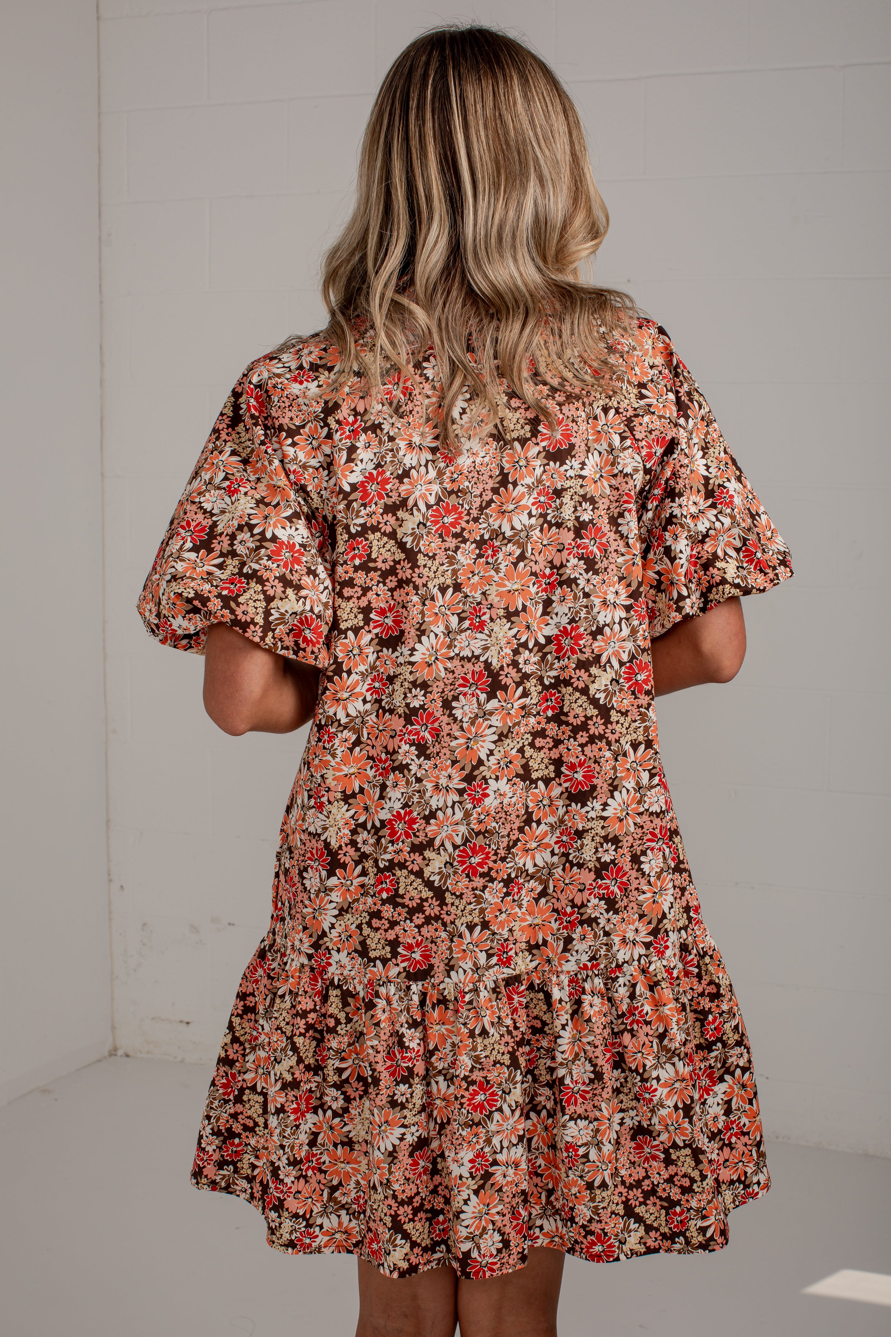 A woman viewed from behind wearing a short-sleeve floral dress with puff sleeves and a tiered skirt, featuring a dense print of red, orange, and white flowers on a dark background.