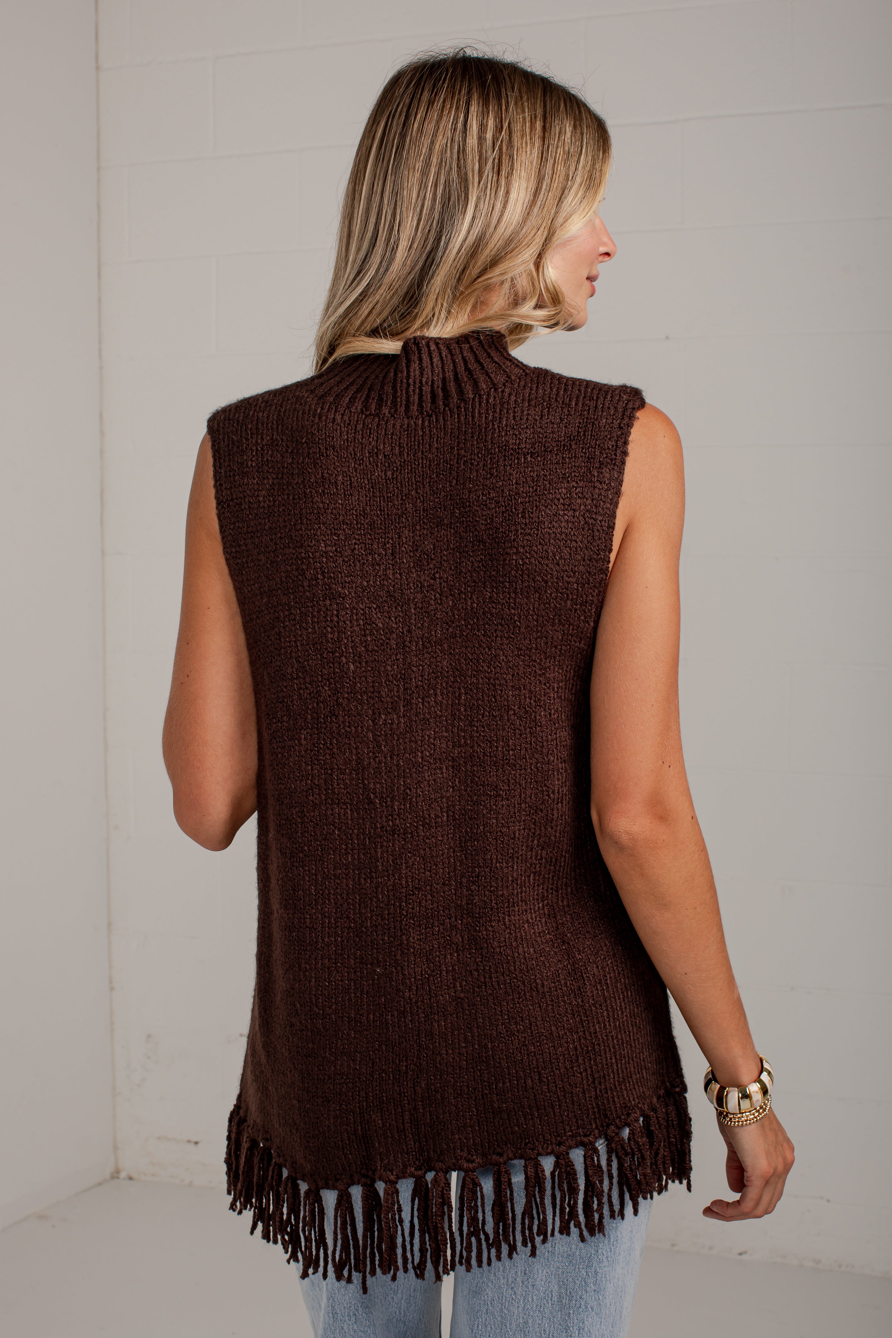 A woman shown from the back wearing a sleeveless, ribbed knit turtleneck vest in dark brown with fringe trim along the hem, paired with light blue jeans against a white studio wall.