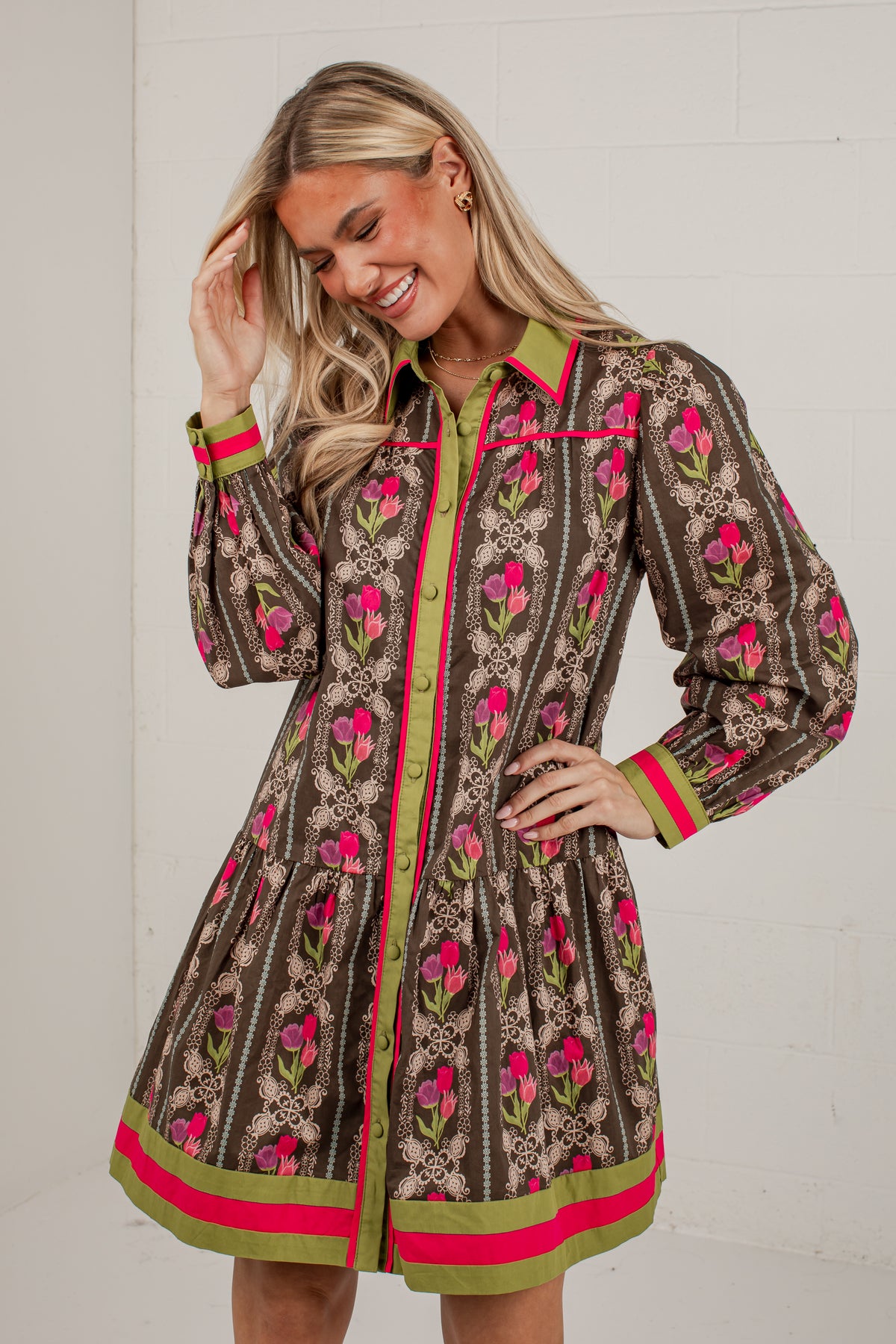 A woman modeling a brown long-sleeve mini dress with a pink and green floral print, featuring a green collar, pink and green trim, and a button-up front.