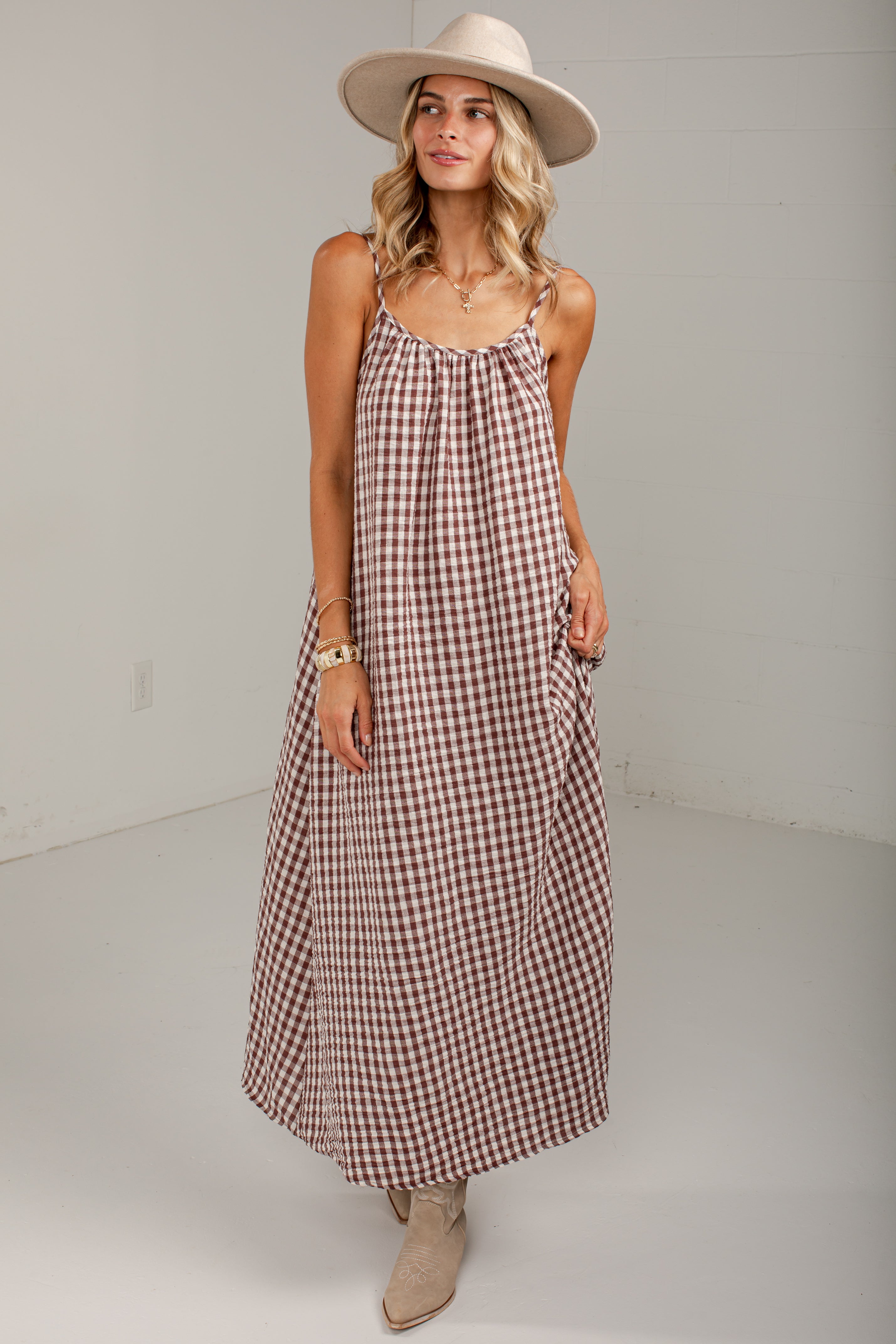 A woman modeling a sleeveless, midi-length gingham dress in brown and white. She is wearing a wide-brimmed beige hat, a gold chain necklace, and beige ankle boots, standing against a plain light gray studio background.