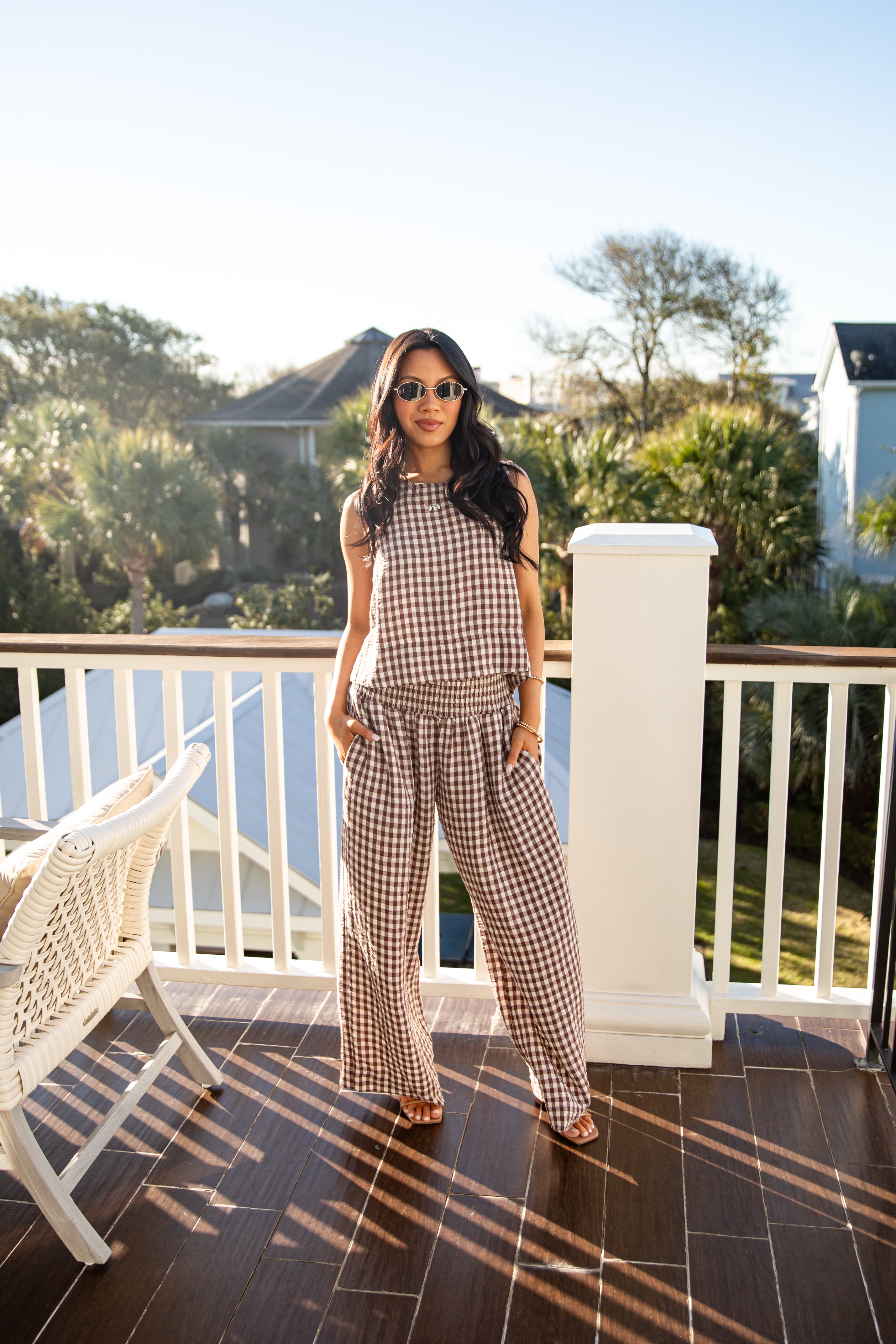 Woman in a checkered outfit standing on a balcony with trees and a building in the background.