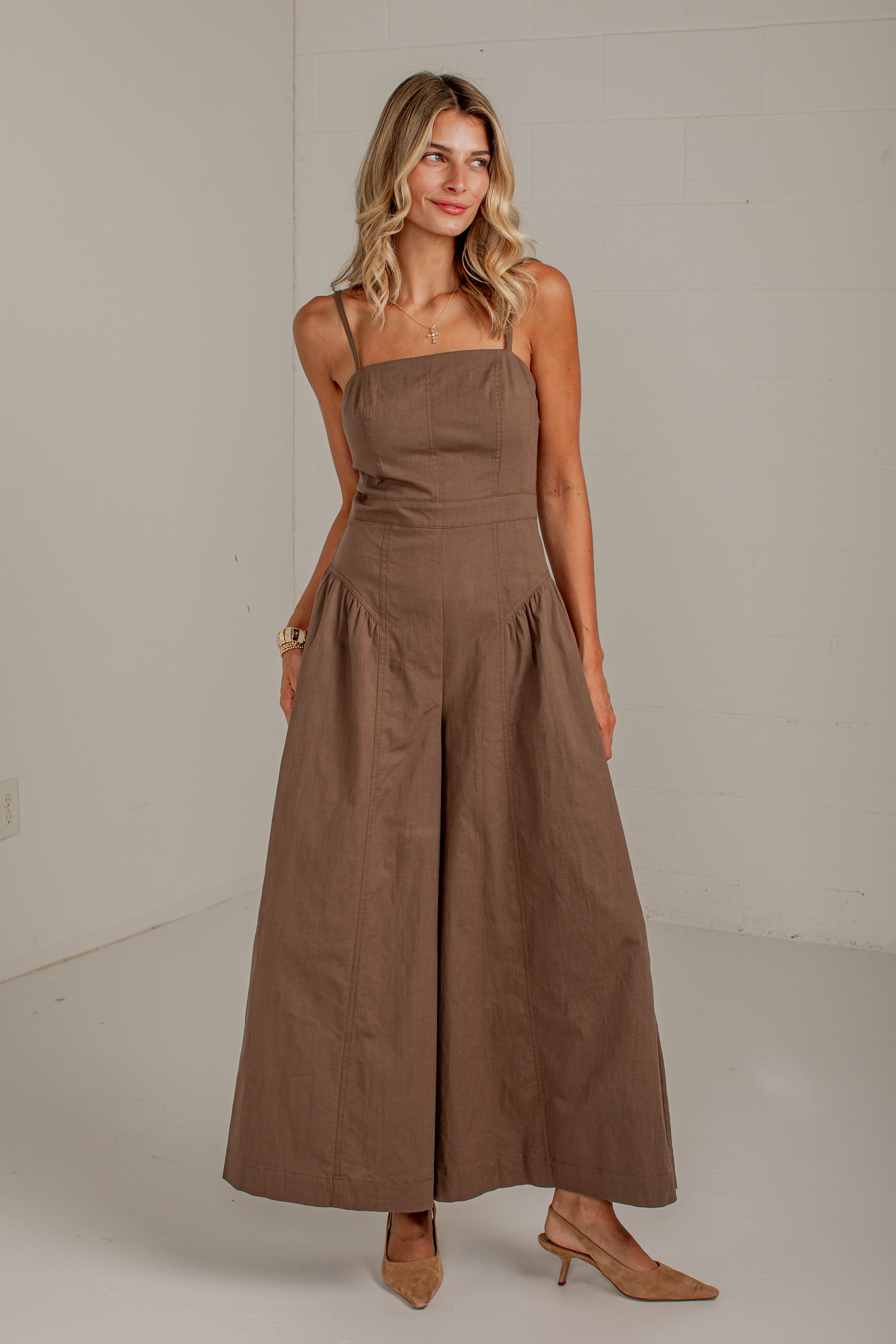 A woman modeling a brown wide-leg jumpsuit with thin straps and a gathered waist, standing against a plain light gray studio wall.