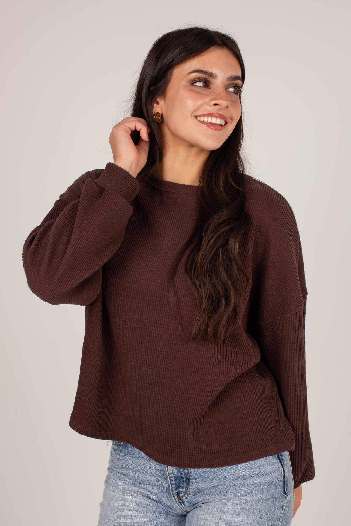 A woman wearing a dark brown ribbed knit sweater and light blue jeans, posing against a neutral light gray background.