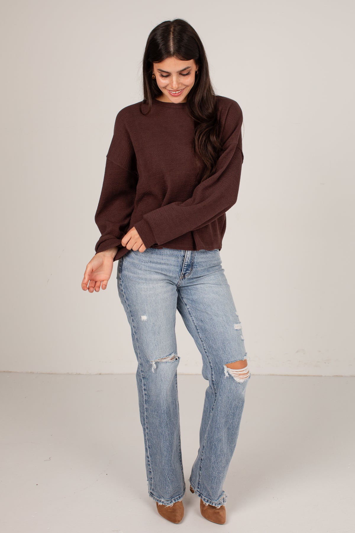 A woman modeling a dark brown long-sleeve knit sweater and light-wash distressed jeans against a plain light gray studio background.