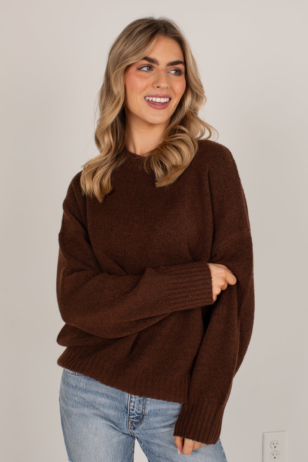 A woman wearing a cozy, oversized dark brown knit sweater and light blue jeans, standing against a plain light gray wall.