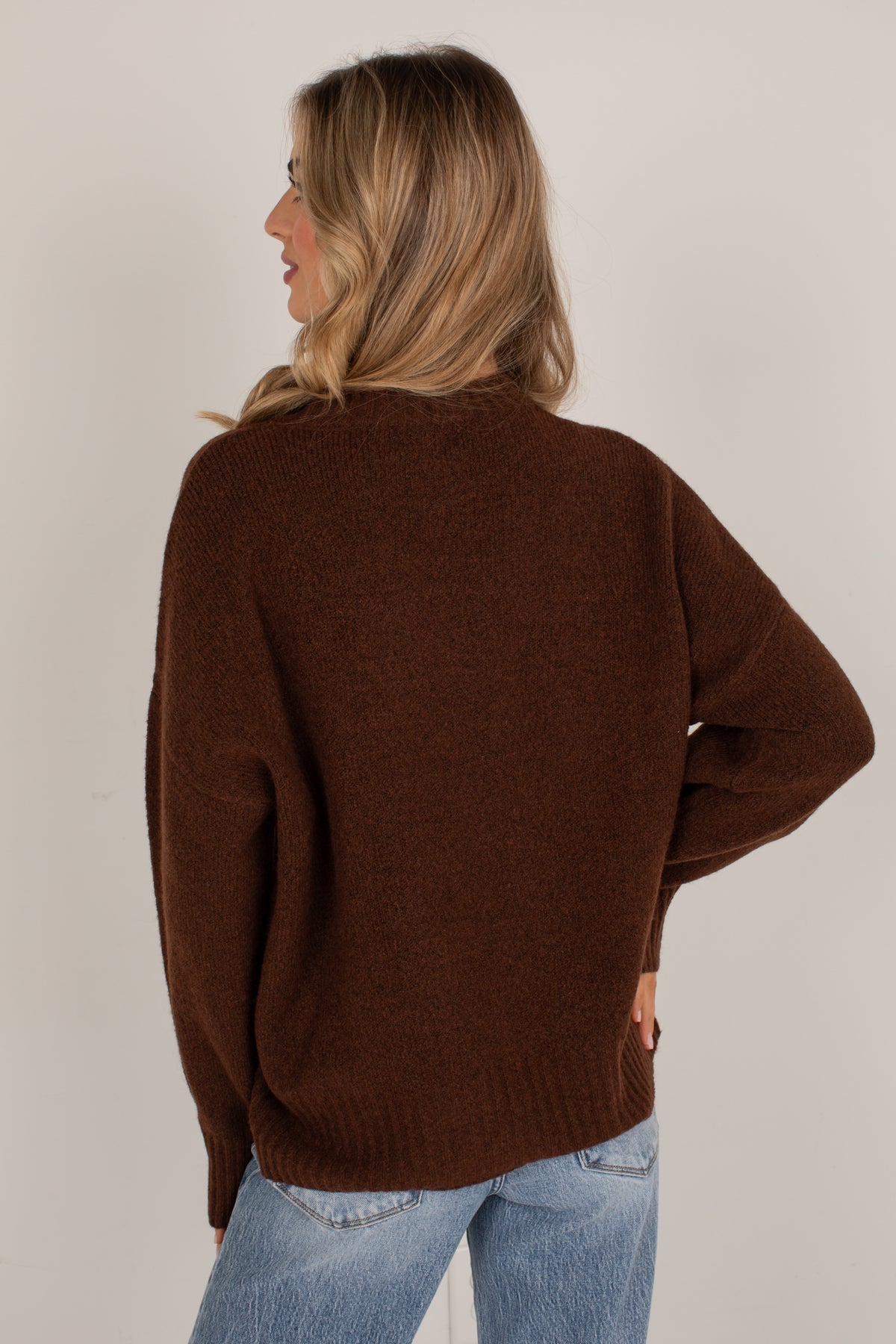 A woman shown from the back wearing an oversized brown knit sweater and light blue jeans, photographed against a plain light gray studio background.