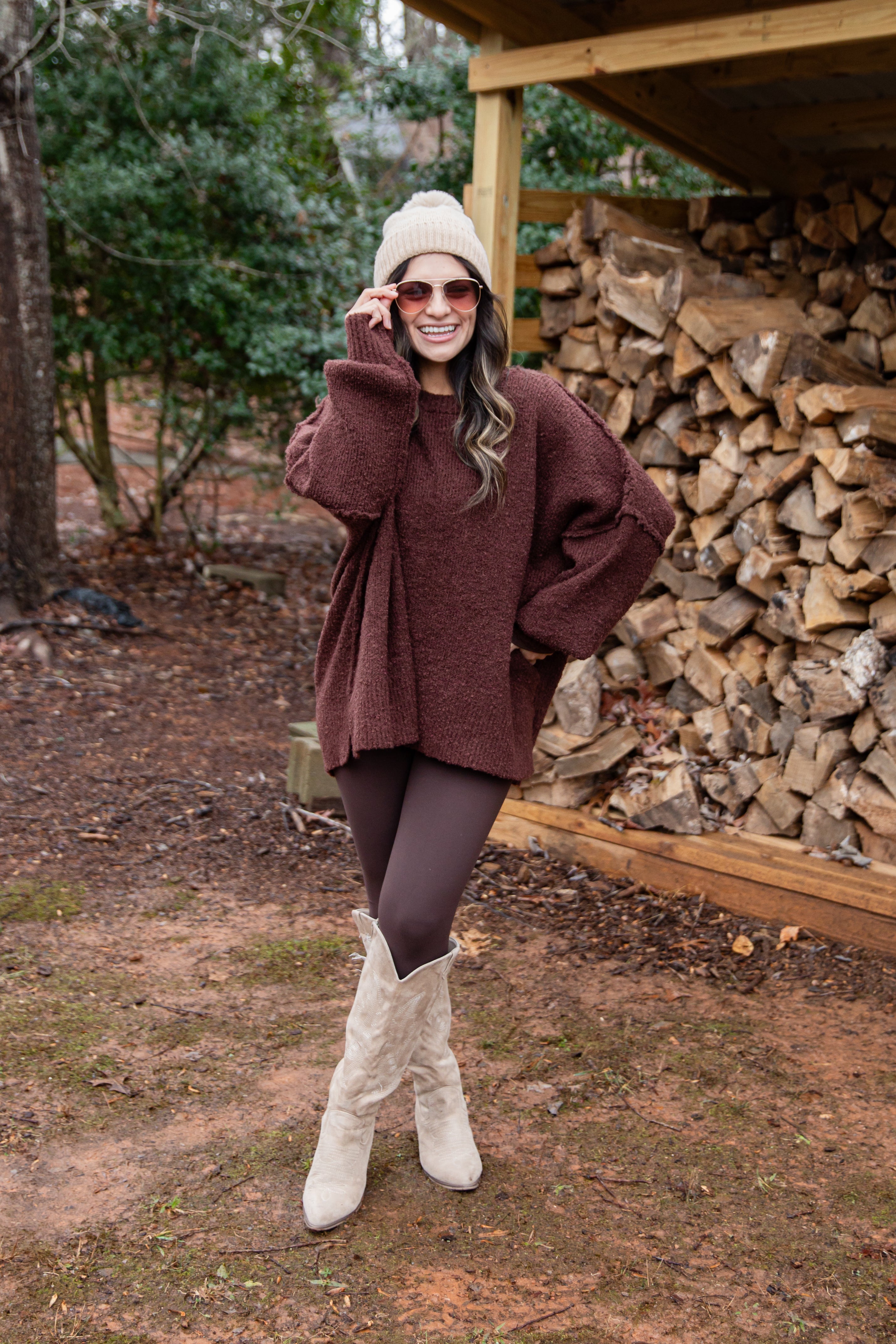 Woman in a brown sweater and white boots standing in front of stacked firewood.